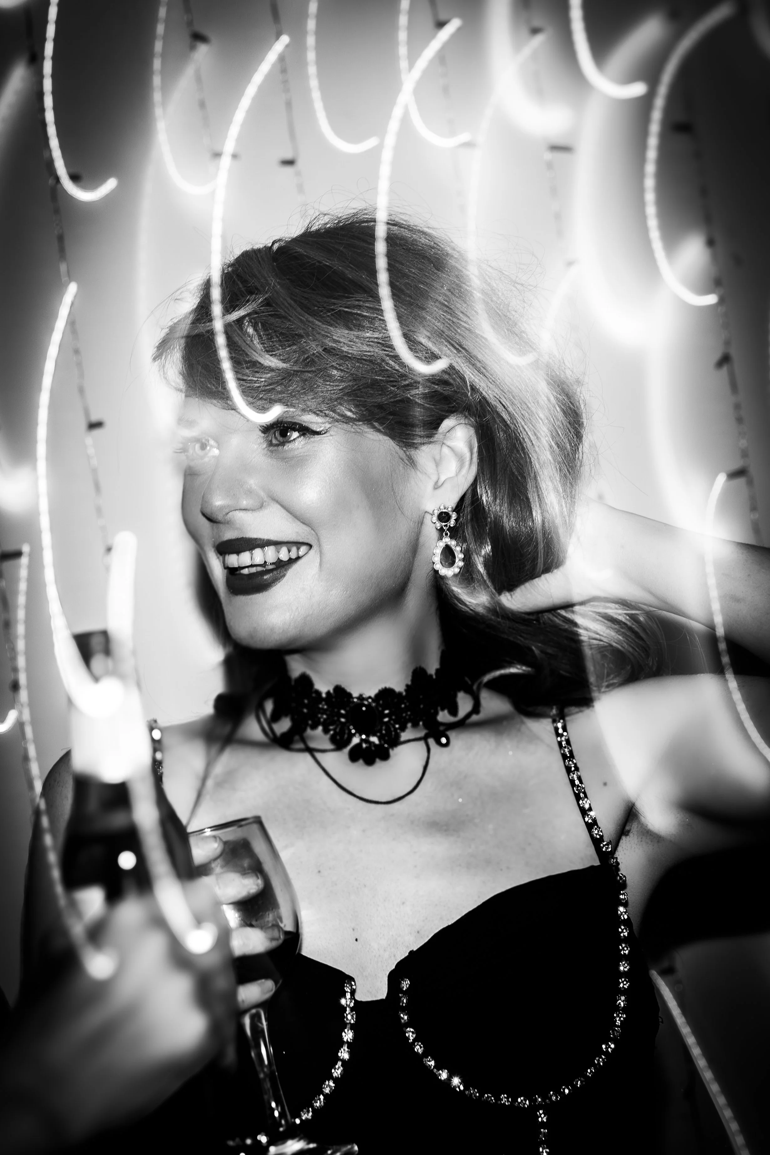 A woman with short hair and jewelry, smiling and holding a glass of wine, at a party or celebration with neon lights in the background.