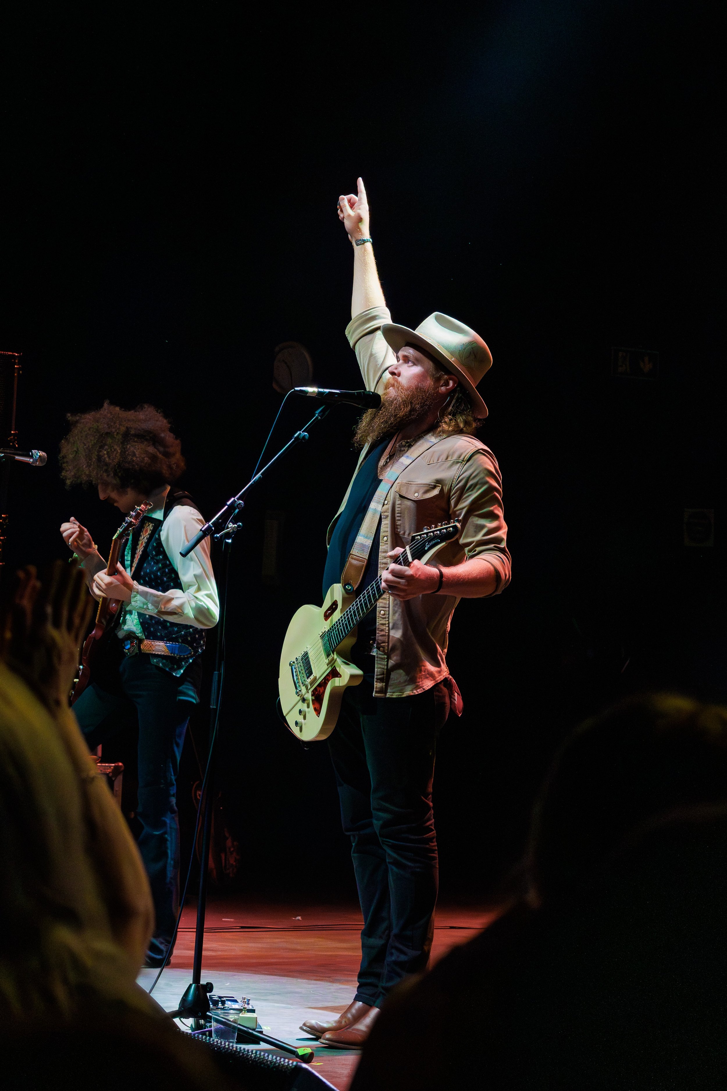 A man with a beard wearing a hat and beige jacket playing an electric guitar on stage, with a person with curly hair also playing an instrument beside him. The stage is dimly lit with a dark background.