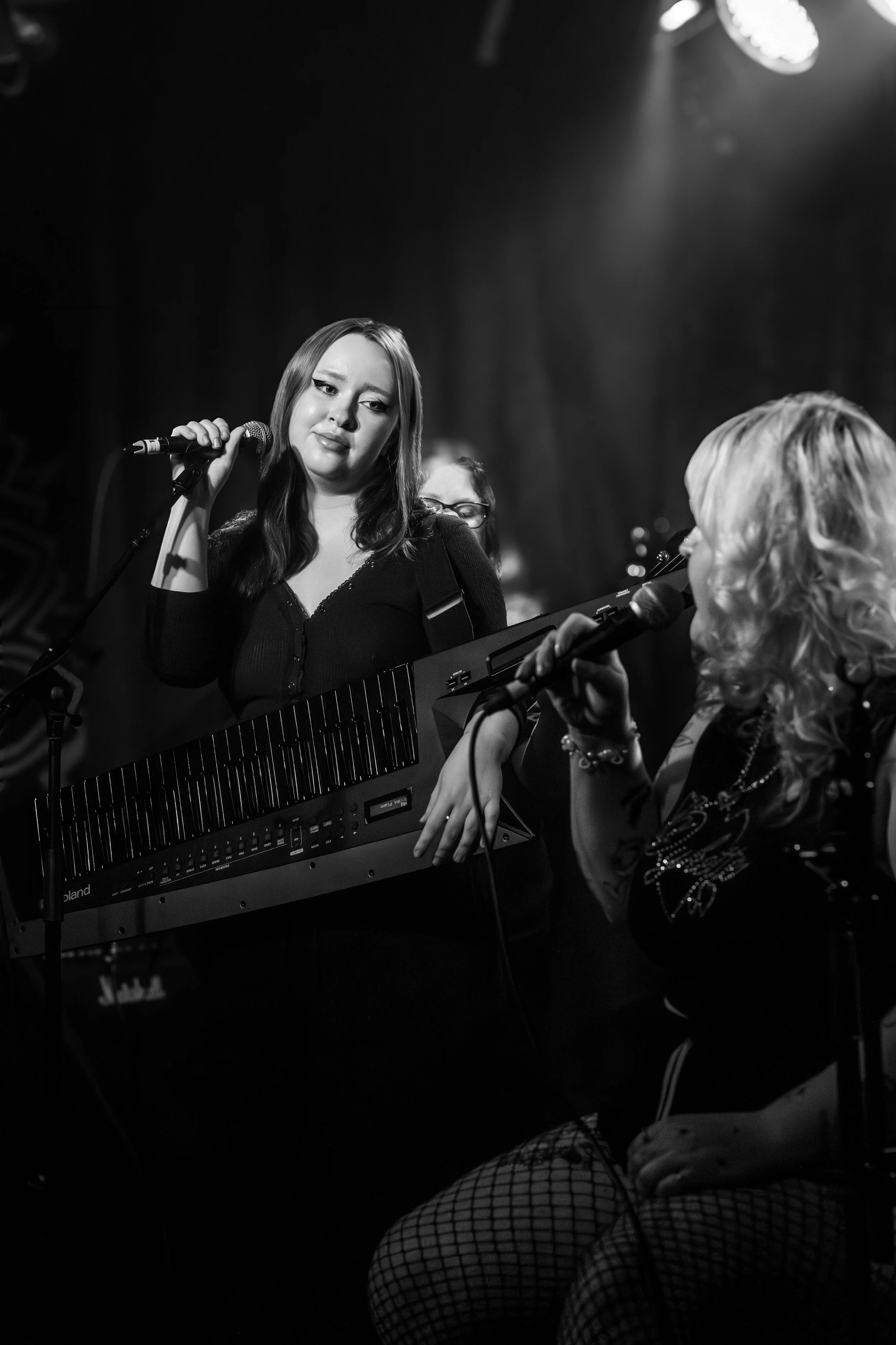 Two women singing into microphones on a dimly lit stage, one holding a keyboard and the other in fishnet stockings.