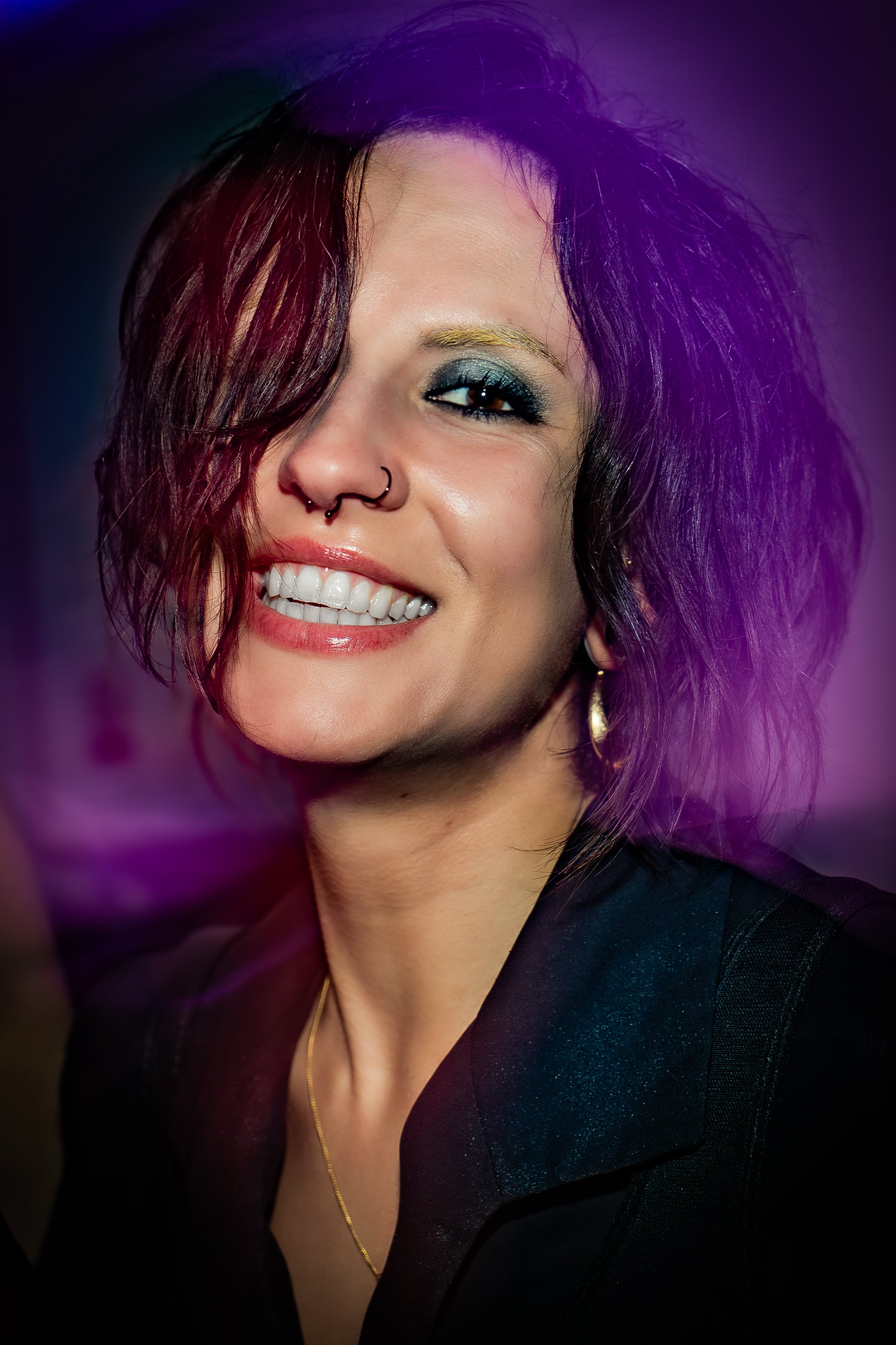 Close-up of a woman with dark, wavy hair, makeup, gold earrings, a nose ring, wearing a black outfit, smiling in low light with purple stage lighting.