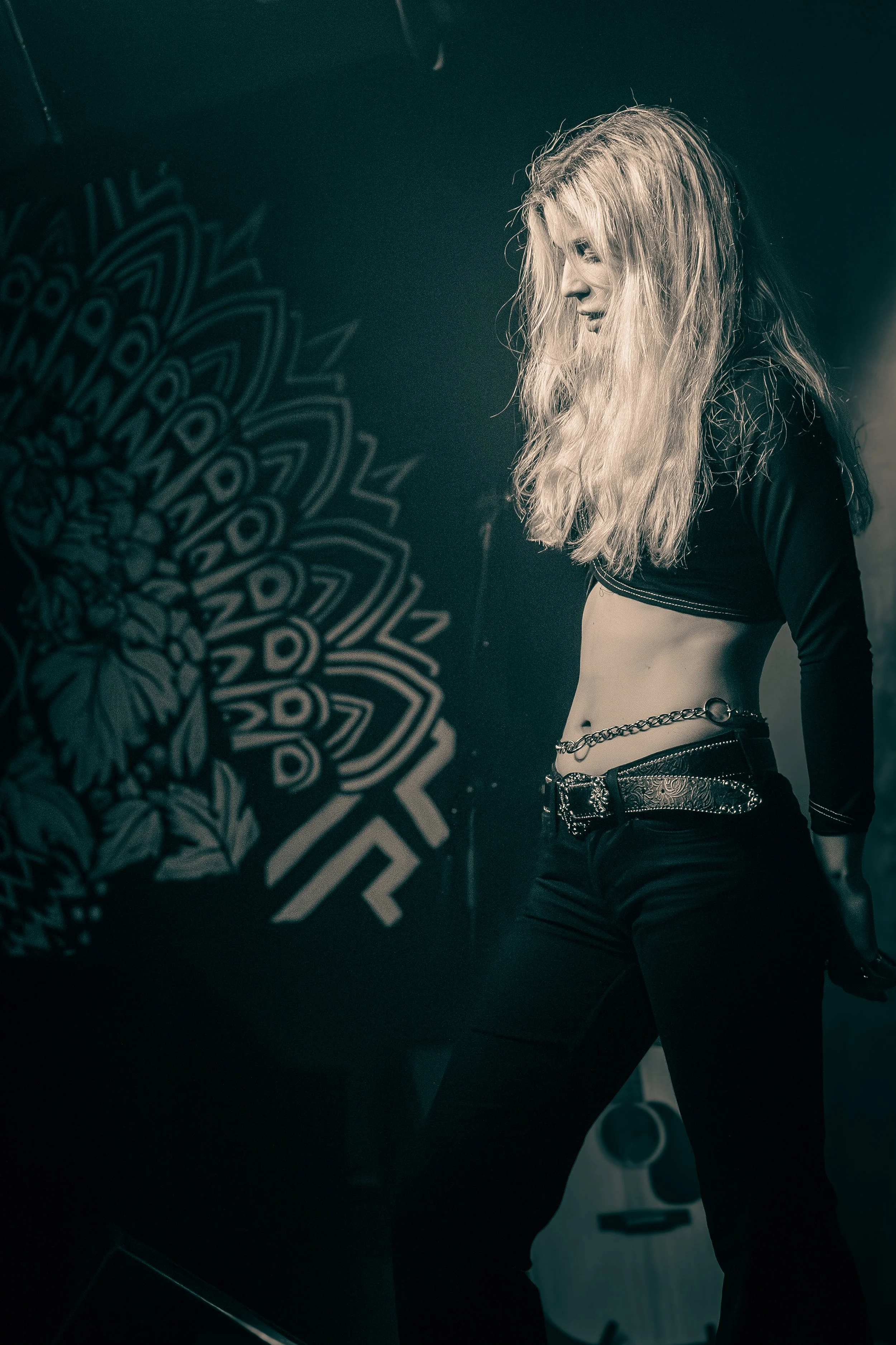 Black and white photo of a woman with long, wavy blonde hair, wearing a cropped black top, black pants, and a decorative belt, standing in front of a patterned wall.