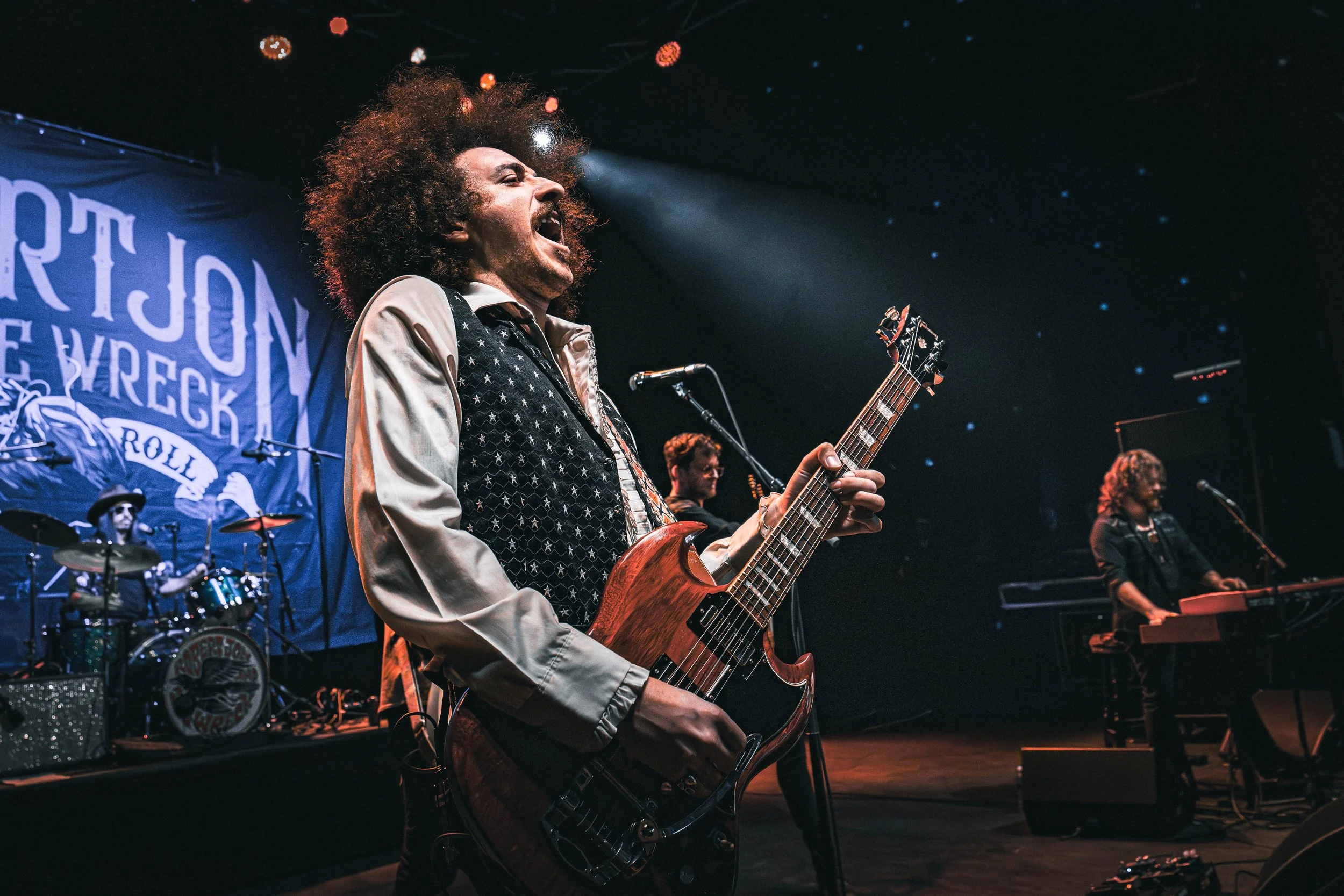 A male musician with curly hair passionately singing and playing an electric guitar on stage during a live performance, with a drum set and other band members visible in the background.