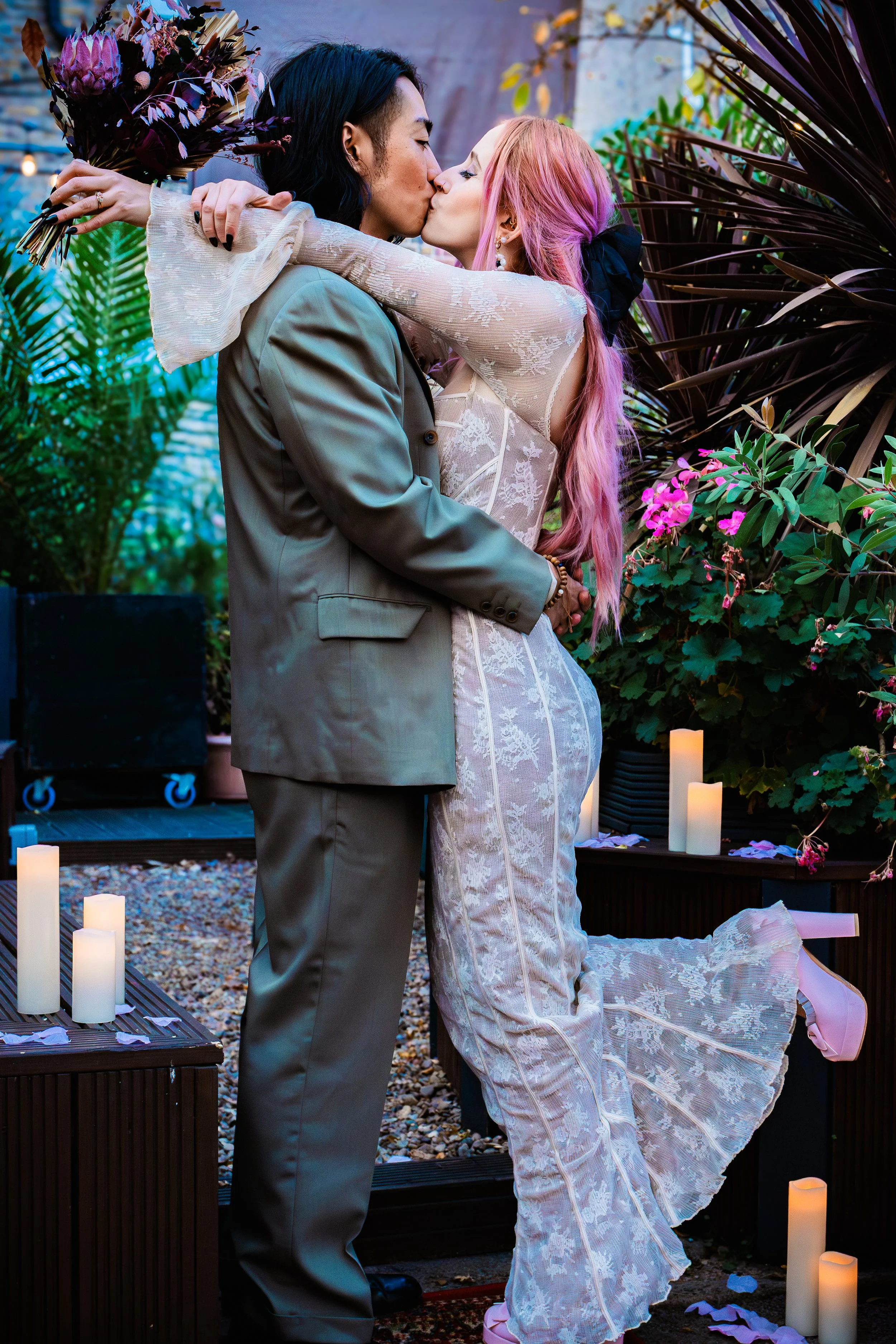 A couple sharing a kiss at their wedding, with the bride raising one leg. The bride has pink hair and is wearing a lace dress, while the groom is in a gray suit. They are surrounded by greenery, candles, and flowers.