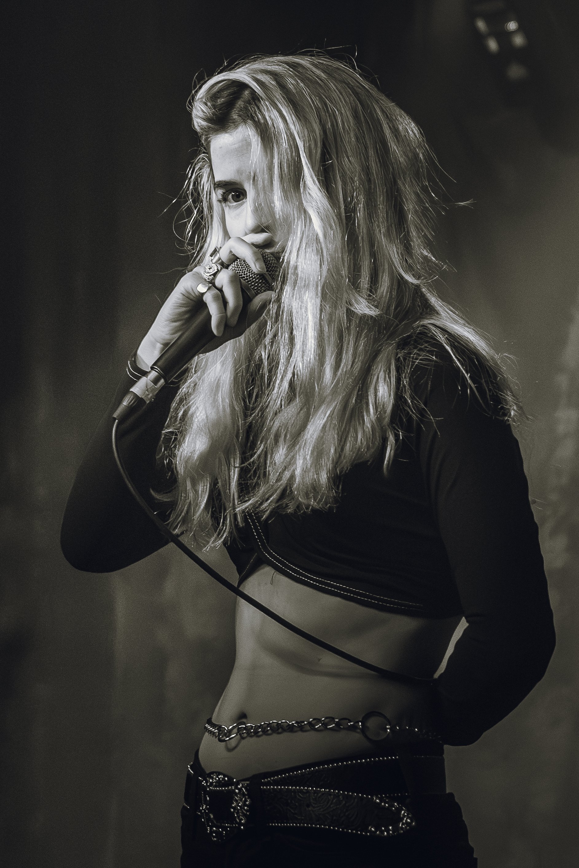A female singer with long, wavy hair performs on stage, holding a microphone close to her lips and looking into the camera.