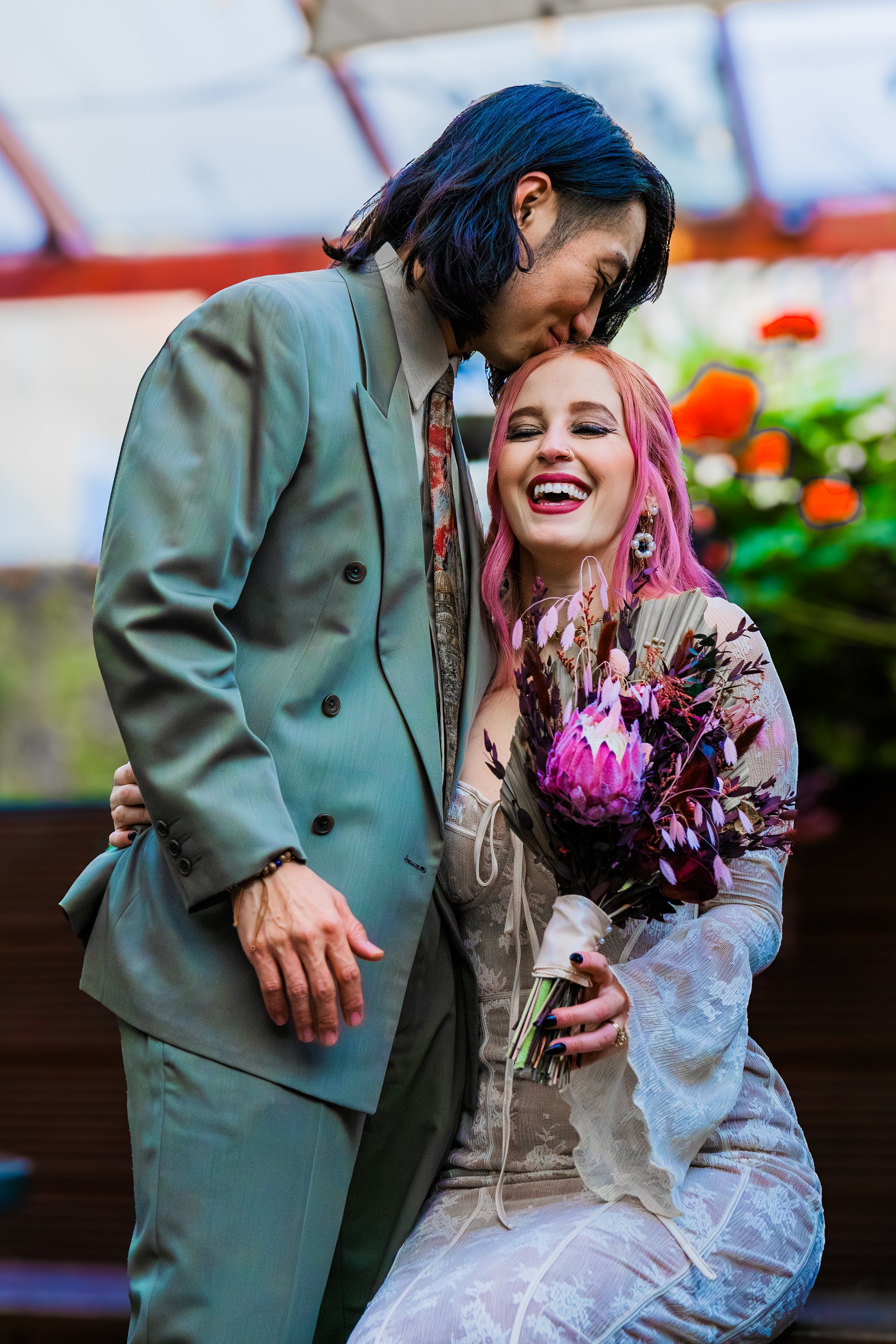 A man with long black hair and a woman with pink hair share a joyful moment, the man kissing the woman's forehead while she holds a bouquet of flowers and laughs.
