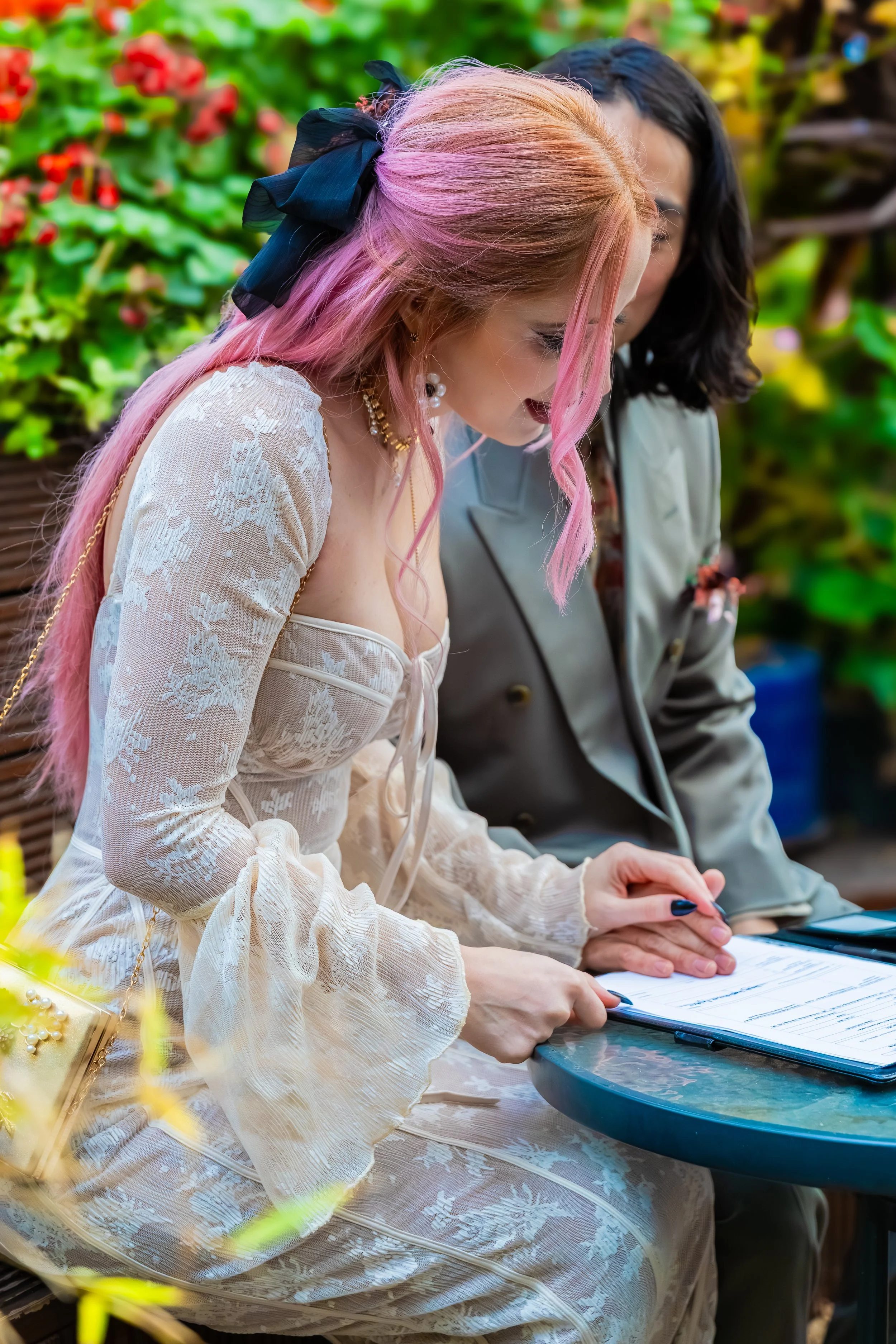 Two women sitting at a table outdoors, looking at a clipboard, with one woman having pink hair and wearing a lace cream-colored dress, and the other woman having dark hair and wearing a gray blazer.