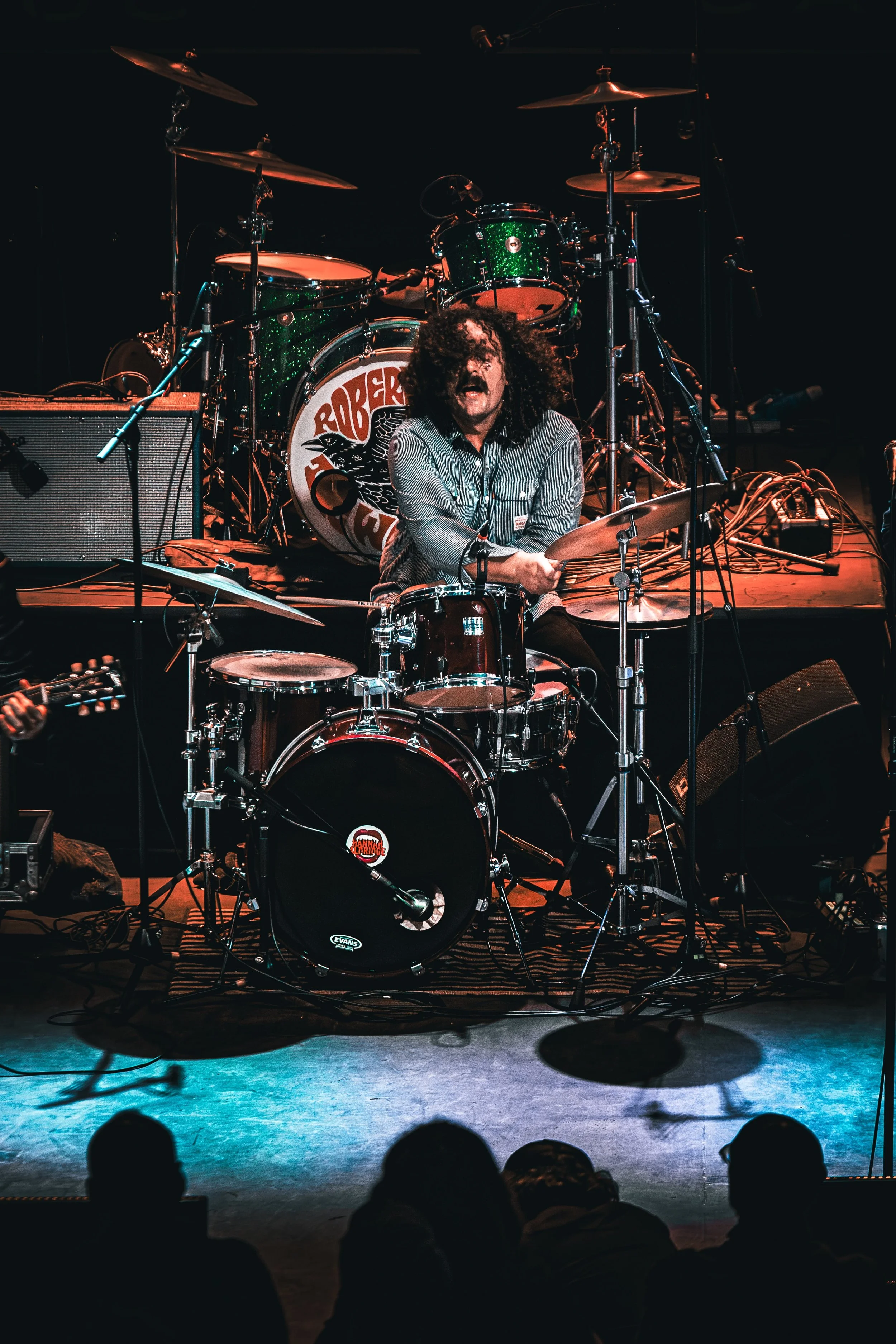 A musician with curly hair and a mustache is playing drums on stage with a green sparkly drum set. The drum set has a bass drum with a Roberta Flack logo. Audience members are visible in silhouette in the foreground.