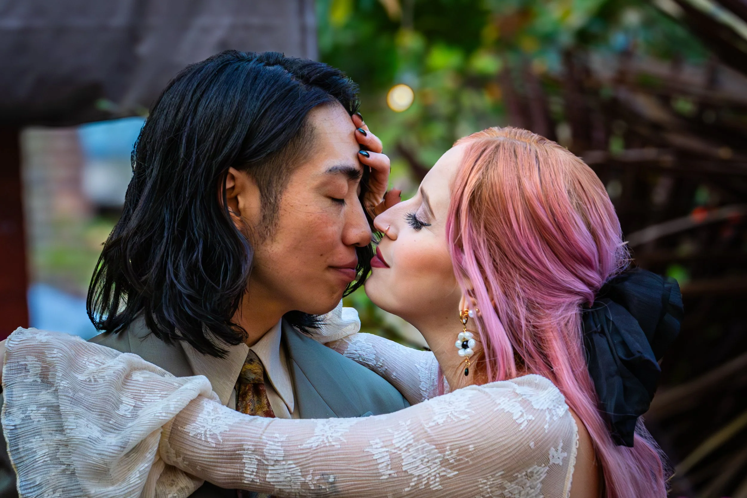 A man and woman are about to kiss, eyes closed, leaning in close. The man has long black hair, and the woman has pink hair and is wearing large earrings and makeup. They are outdoors with blurred greenery in the background.