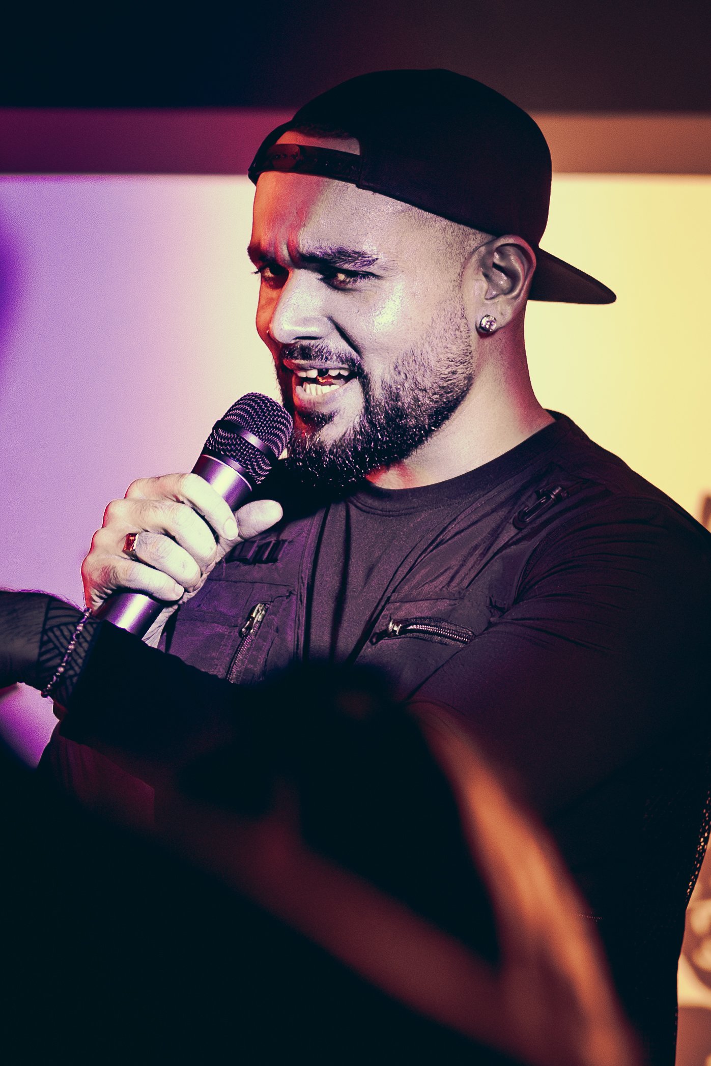 A man with a beard and earrings wearing a black cap backwards and black shirt, speaking into a microphone on stage, with colorful lights in the background.