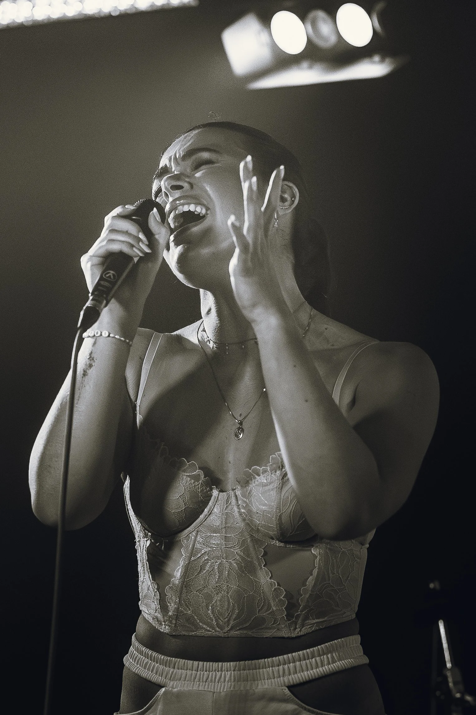 A woman singing passionately with a microphone, wearing a lace top and athletic pants, in a concert setting with stage lights overhead.