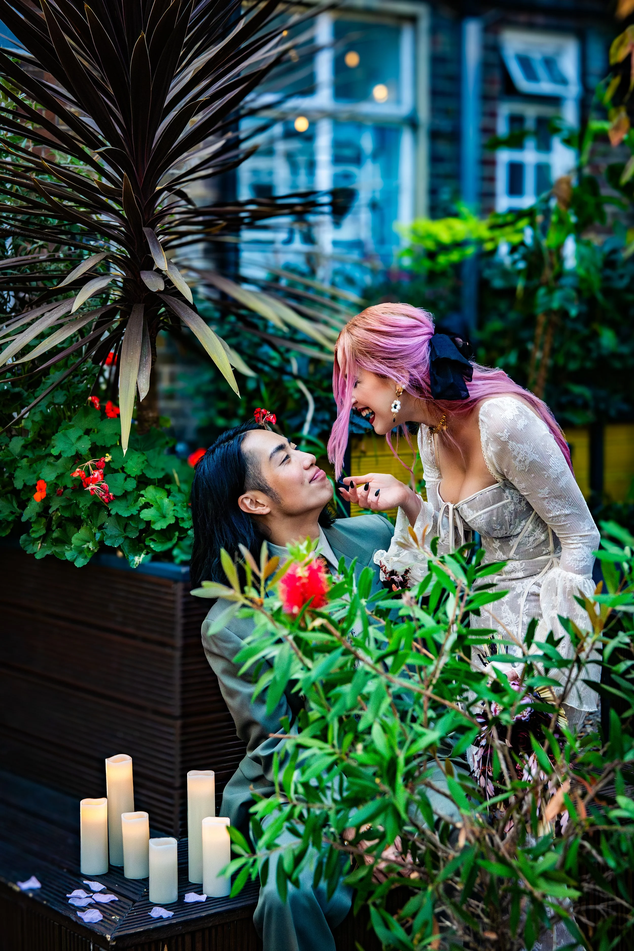 A couple dressed in wedding attire in a lush green garden surrounded by plants and flowers. The woman with pink hair is leaning towards the man with long dark hair, smiling and playfully touching his chin. There are candles on a small table with flower petals scattered nearby.