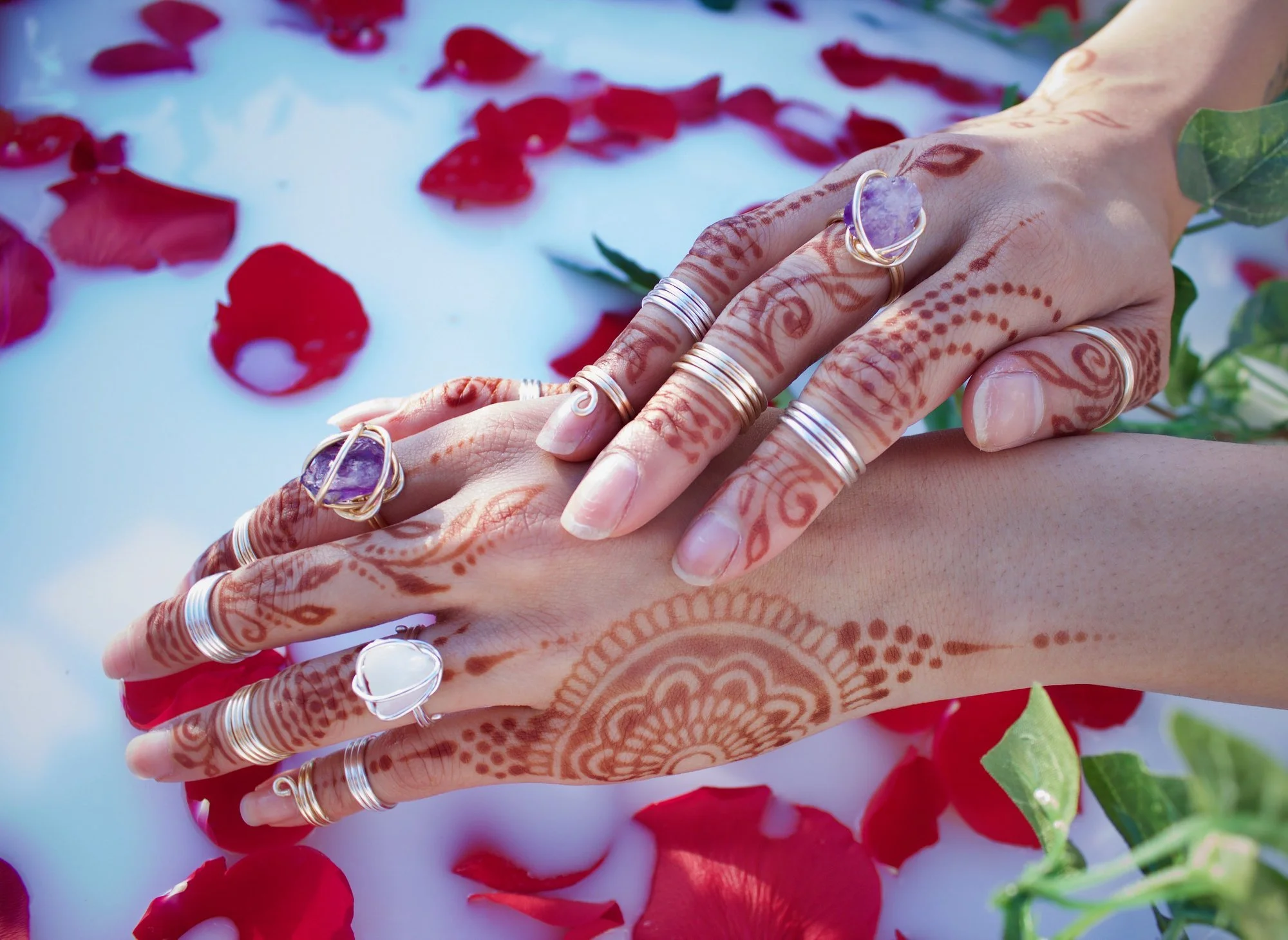 Two hands with intricate henna designs and multiple silver rings, some with purple and white stones, resting on a white surface decorated with rose petals and greenery.