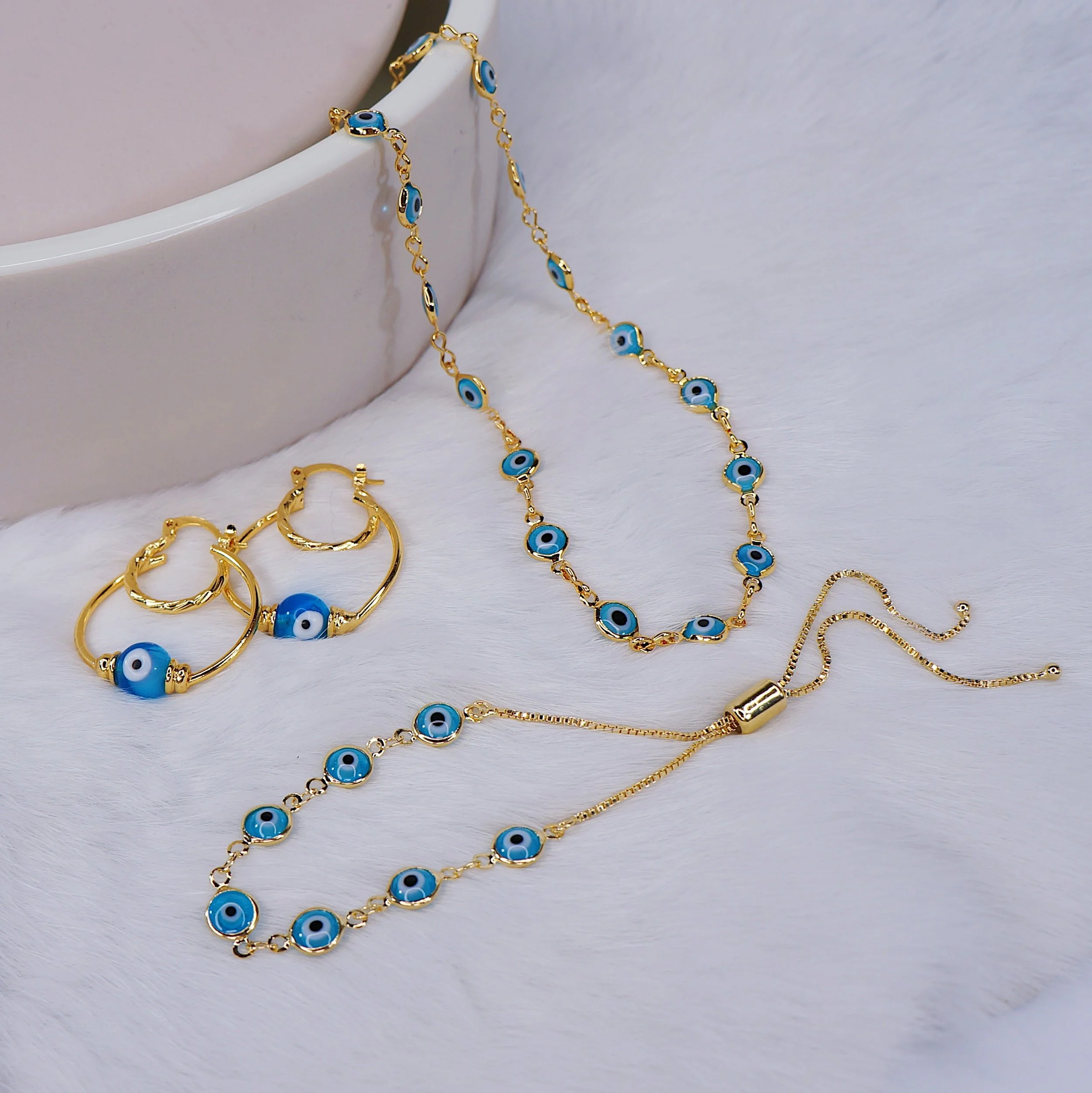 Gold jewelry set with blue evil eye symbols, including a necklace, a bracelet, and a pair of earrings, displayed on a white surface with a ceramic dish in the background.
