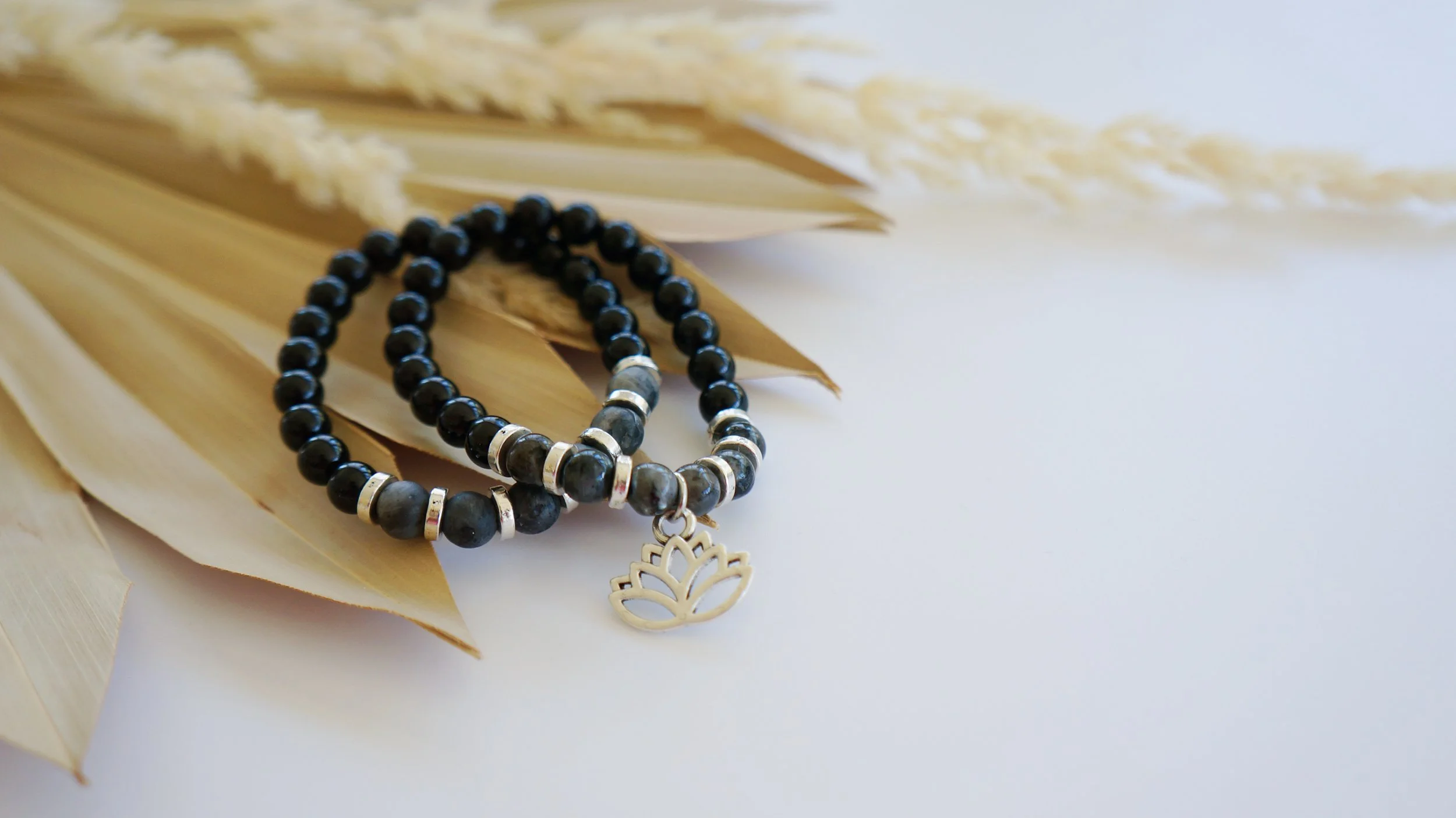 A beaded black bracelet with silver accents and a lotus charm, resting on dried beige palm leaves against a white background.
