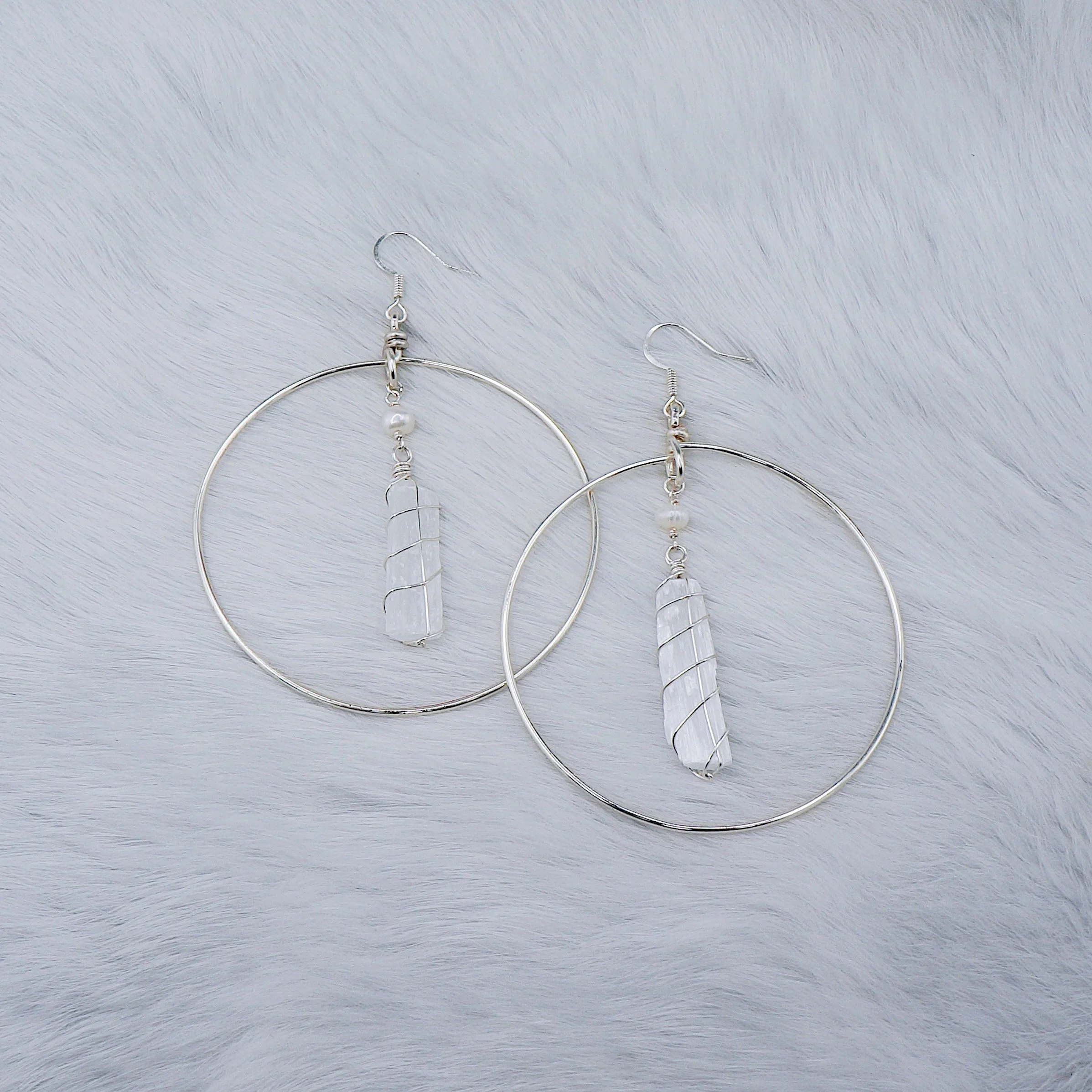 Pair of long silver hoop earrings with spiral wrapped clear stones in the center, laid on a textured white surface.