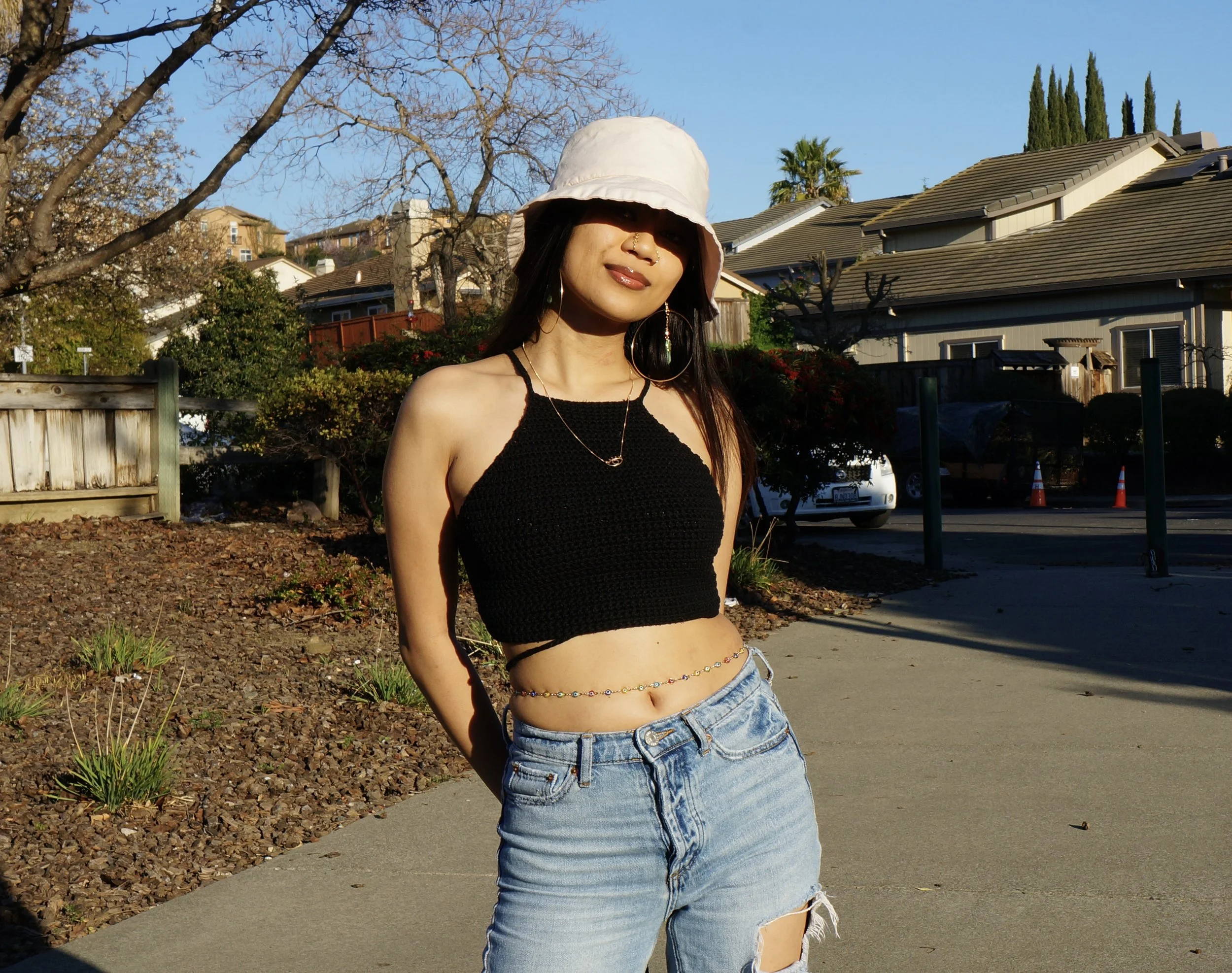 A young woman standing outdoors on a sunny day. She is wearing a white bucket hat, a black crop top, light blue ripped jeans, and has accessories including a necklace, large hoop earrings, and a waist chain. She is smiling with her eyes partially shaded by the hat, with houses, trees, and a clear blue sky in the background.