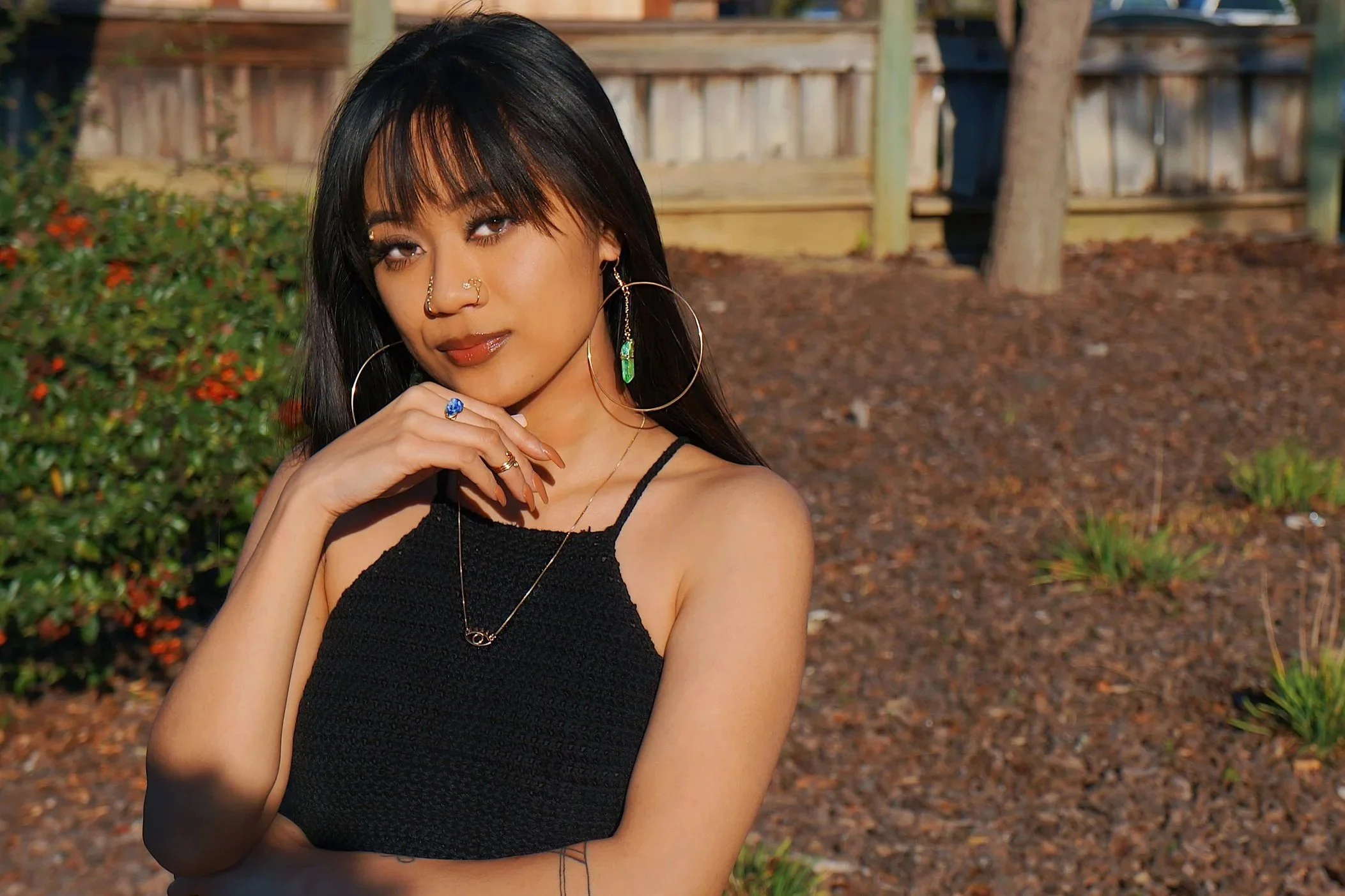 A young woman with dark hair and bangs, wearing a black sleeveless top, multiple rings, hoop earrings, and a necklace, standing outdoors with brown mulch, green plants, and a wooden fence in the background.