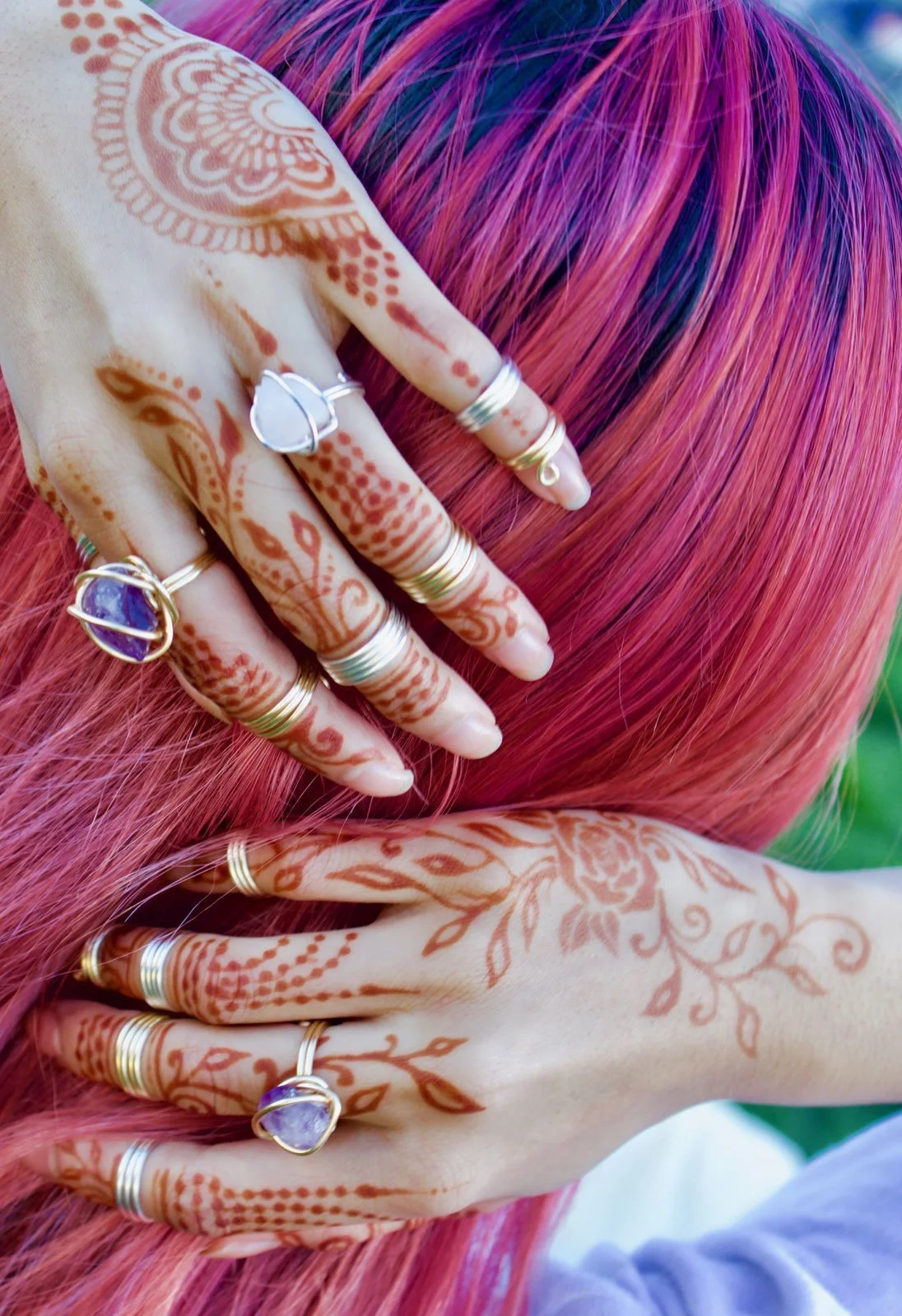 Hands adorned with multiple rings holding vibrant pink hair with purple streaks, featuring henna designs on the hands and fingers.