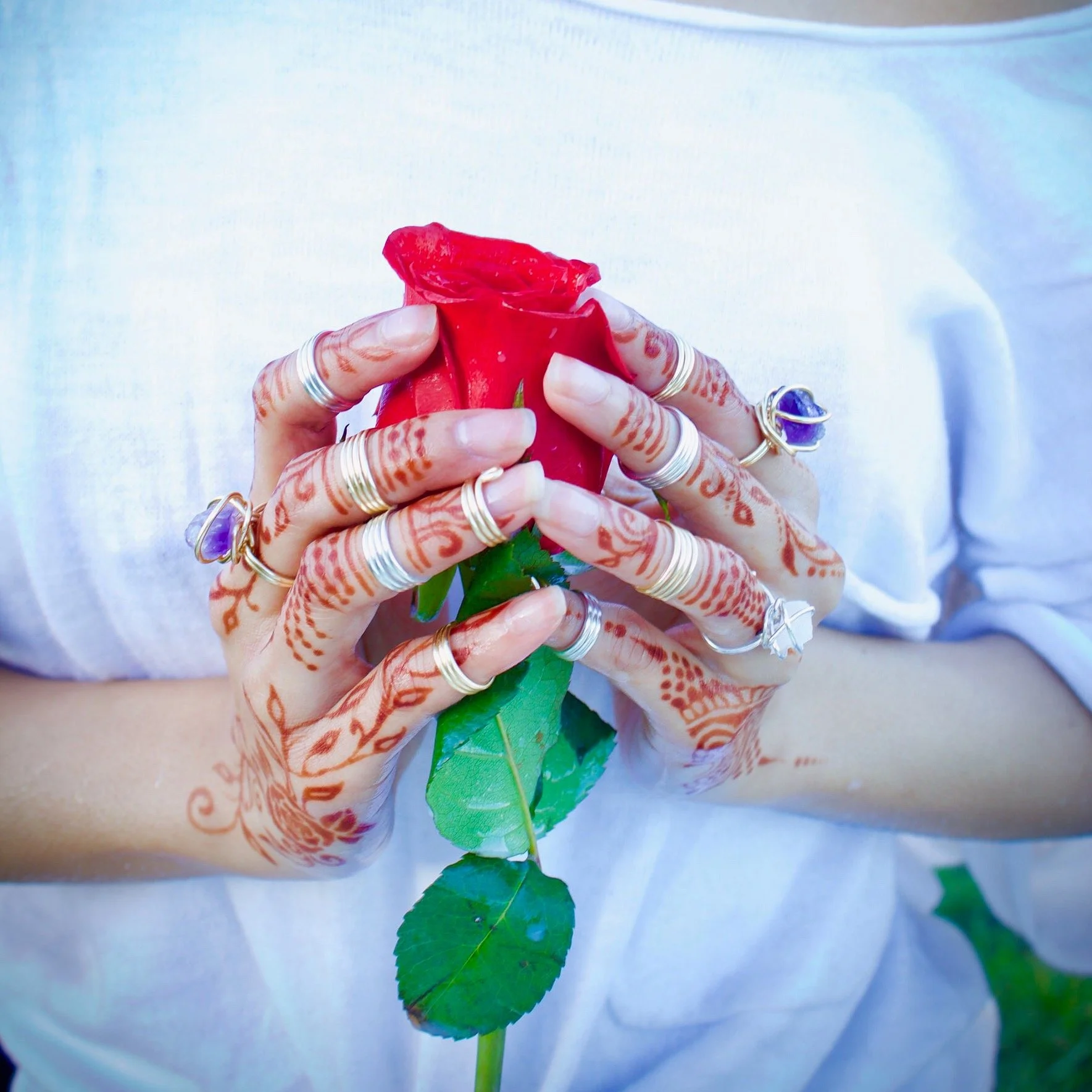 Person holding a red rose with hands decorated with multiple silver rings and henna designs, wearing a white shirt.