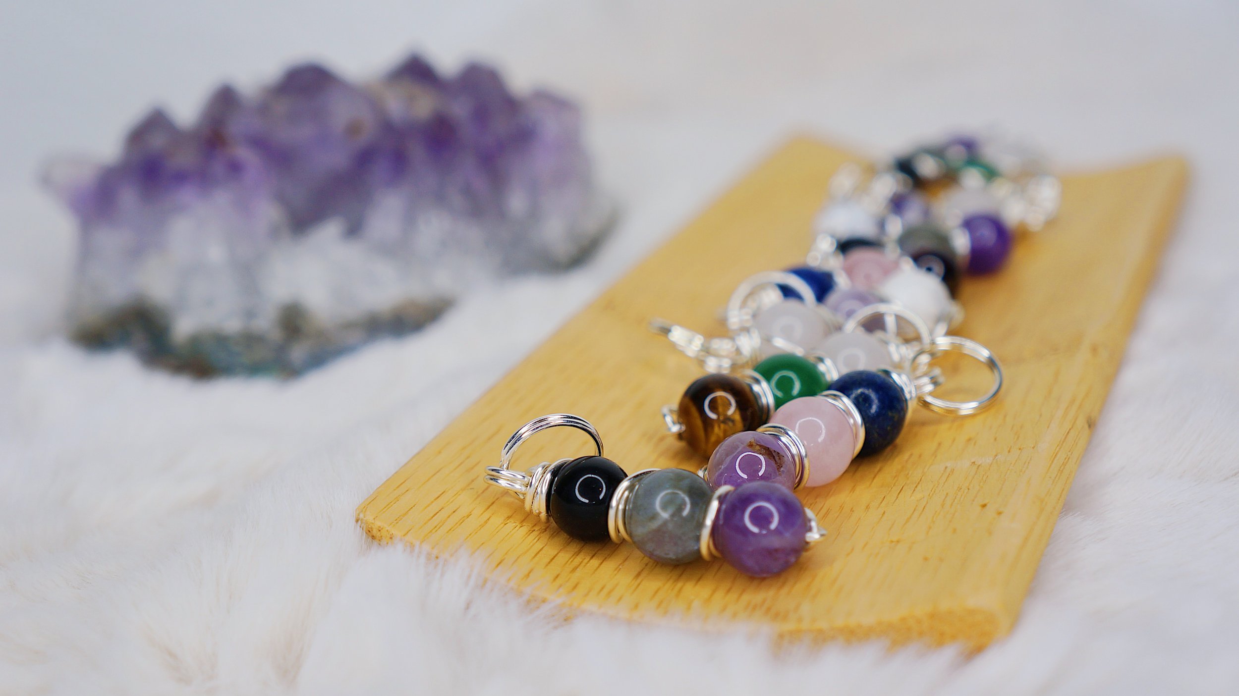 Colorful beaded keychains or charms with silver rings on a wooden surface, with a purple mineral or crystal formation in the background.