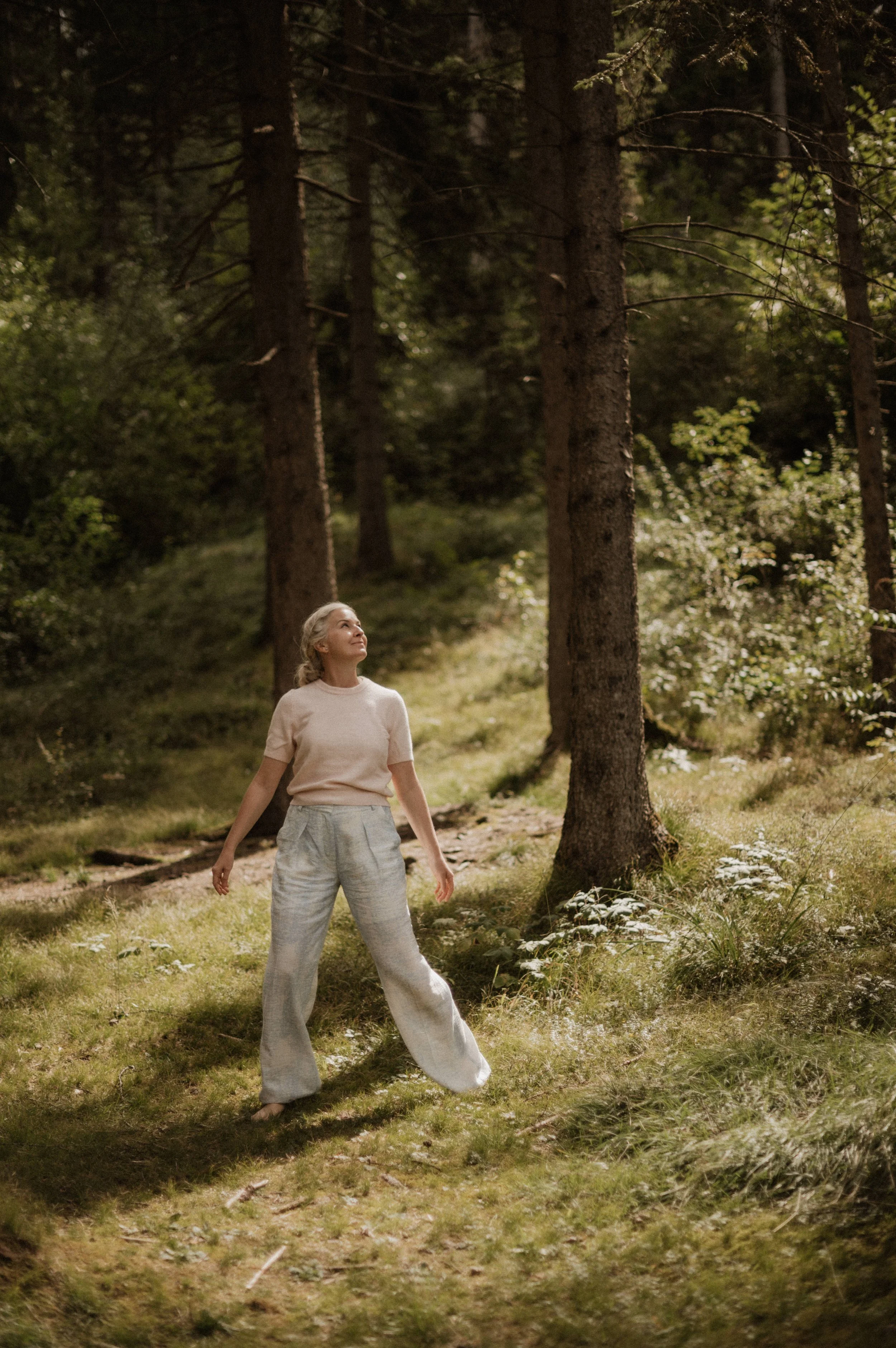 Eine Frau steht in einem sonnendurchfluteten Wald, umgeben von Bäumen, und schaut nach oben.
