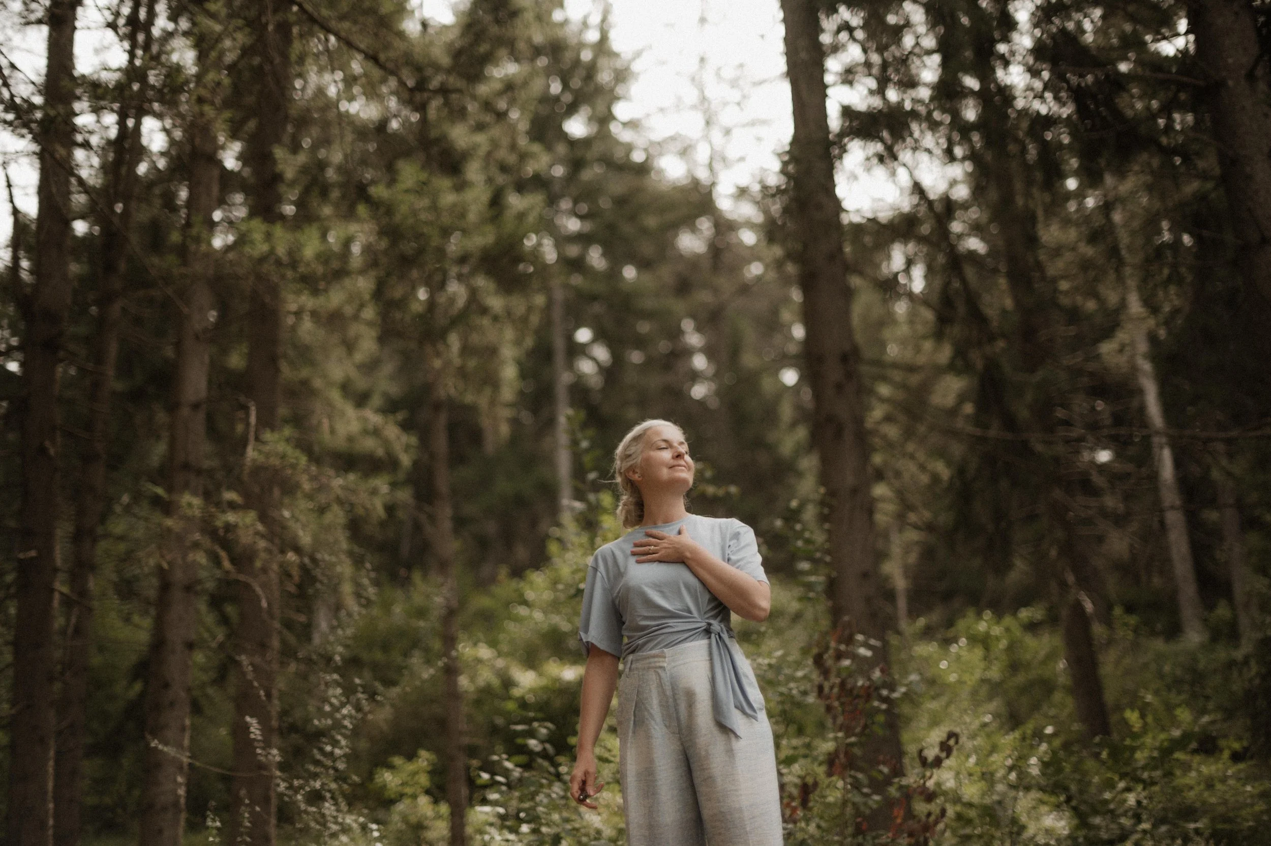 Eine Frau in einem hellblauen Oberteil und grauen Hosen steht in einem grünen Wald, hält eine Hand an die Brust, schaut friedlich nach oben.