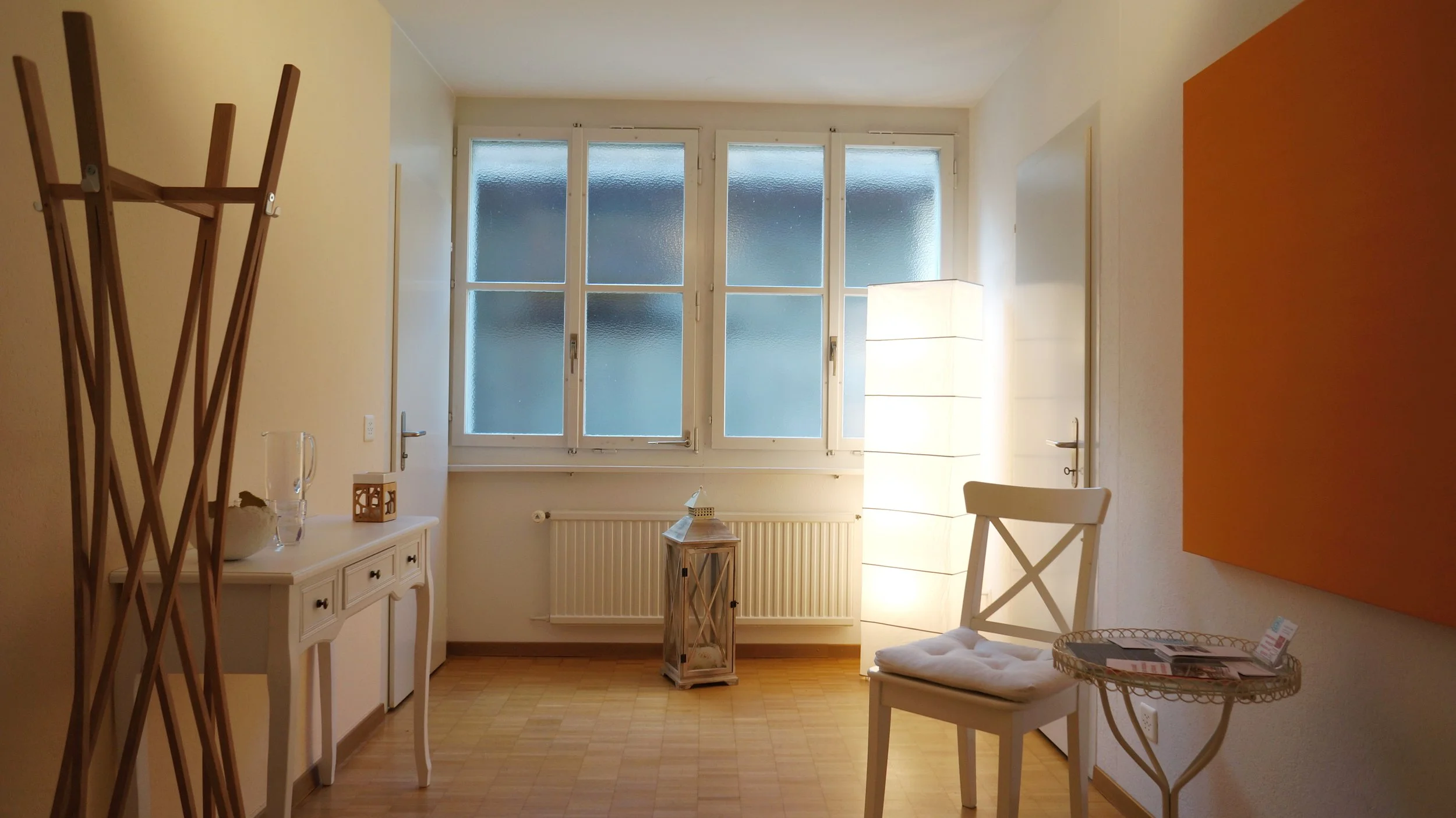 Helles Wohnzimmer mit großen Fenstern, einem weißen Sideboard mit Dekoration, einem weißen Stuhl mit Kissen, einem runden Beistelltisch mit Magazinen, einem großen orangefarbenen Gemälde und einer Stehlampe.