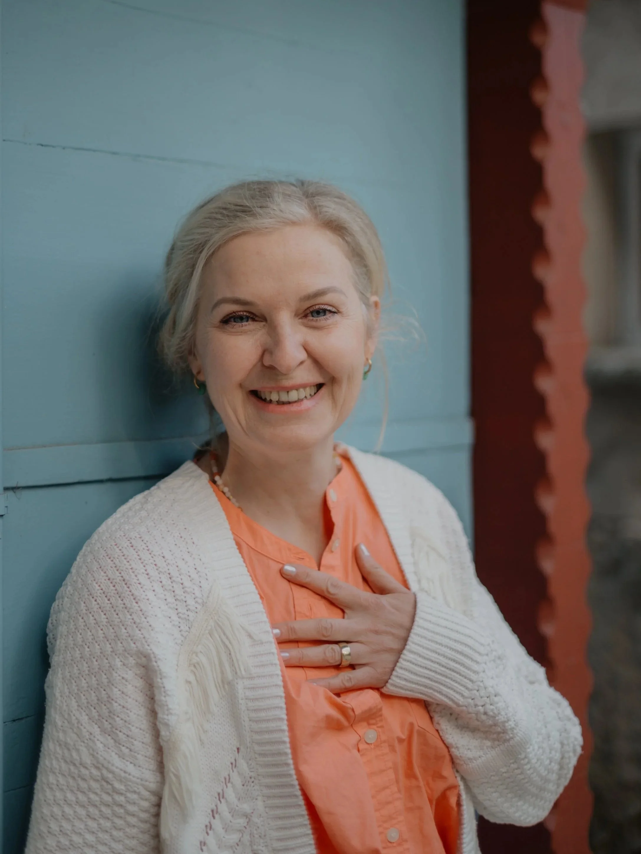 Ein lachender Mann mit blonder, lockiger Frisur trägt ein orangefarbenes Hemd und eine weiße Strickjacke. Sie hält ihre rechte Hand auf die Brust vor sich, trägt eine Ehering, und steht vor einer blauen Wand.