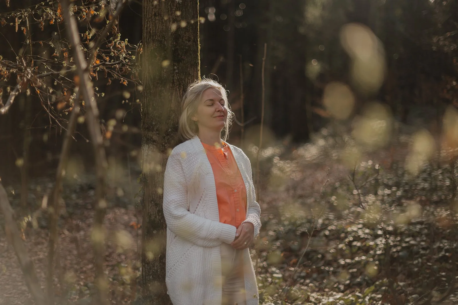Eine Frau entspannt sich im Wald mit geschlossenen Augen, lehnend an einen Baum, während die Sonne durch die Bäume scheint.