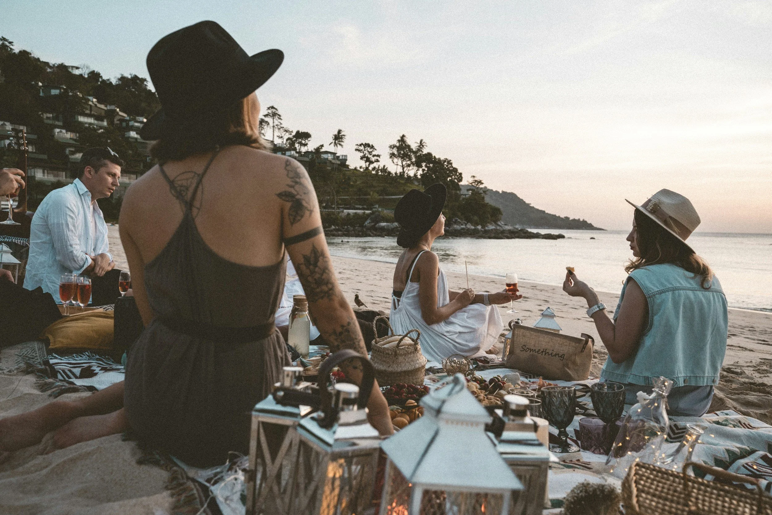 People having a picnic on the beach during sunset, with some sitting on blankets and others standing. They are enjoying drinks and snacks, with lanterns and baskets around them, and the ocean and hillside visible in the background.