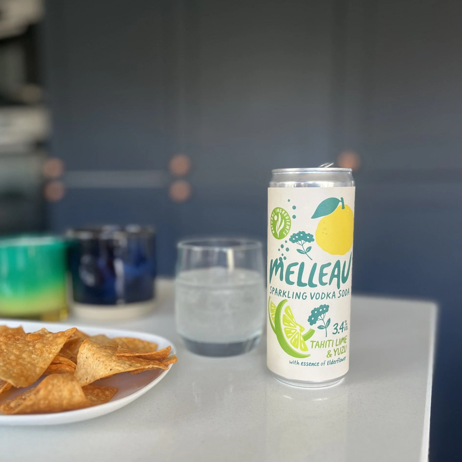 A can of Melleau sparkling vodka soda with lemon and lime flavor, placed on a white surface next to a glass of water and a plate of crisps.