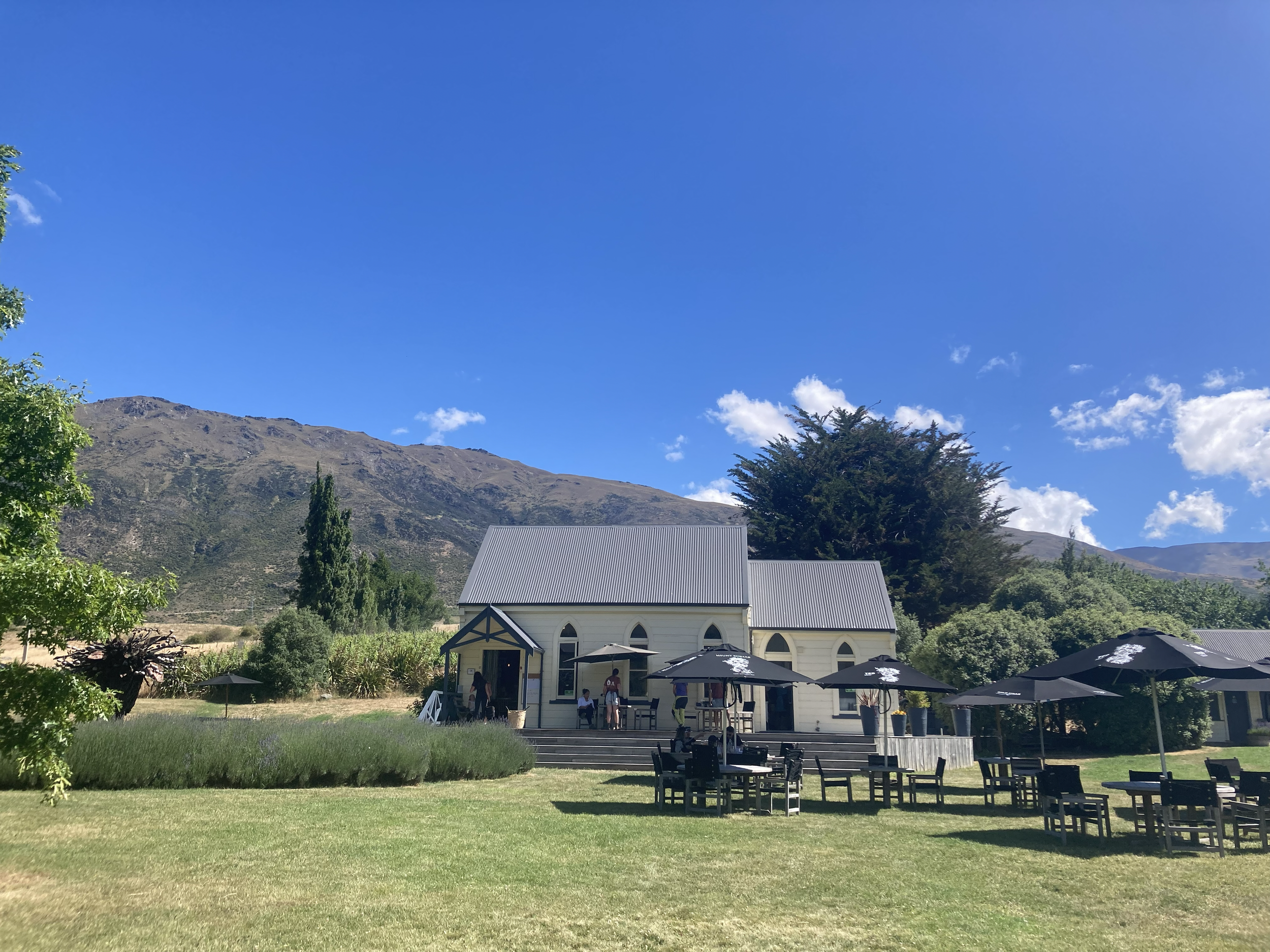 A white church with a gray metal roof and arched windows is set against a mountain landscape under a bright blue sky. In front, there are outdoor tables with black umbrellas and chairs, with some people standing and sitting near the church entrance.