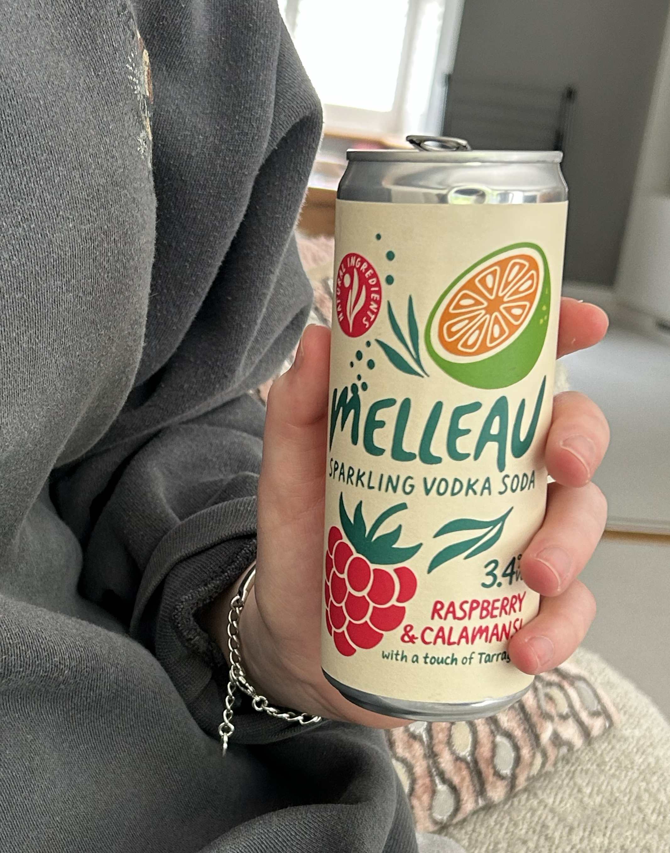A person holding a can of Melleau sparkling vodka soda with raspberry and calamansi flavor, featuring a colorful label with illustrations of a calamansi lime and raspberry.