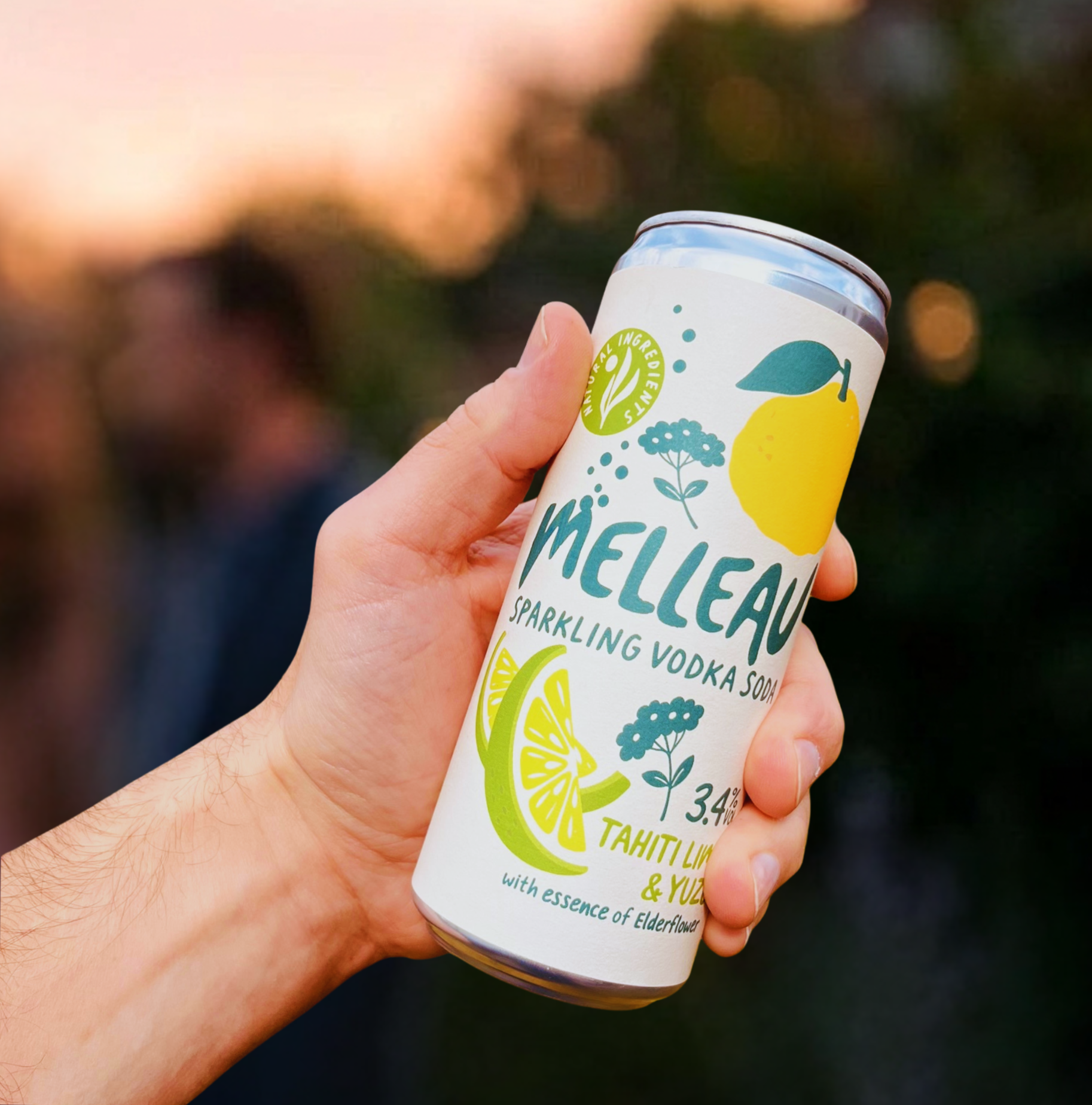 A person's hand holding a can of Melleau sparkling vodka soda with lemon and elderflower flavor, showing a white label with colorful lemon and elderflower illustrations, outdoors during sunset.
