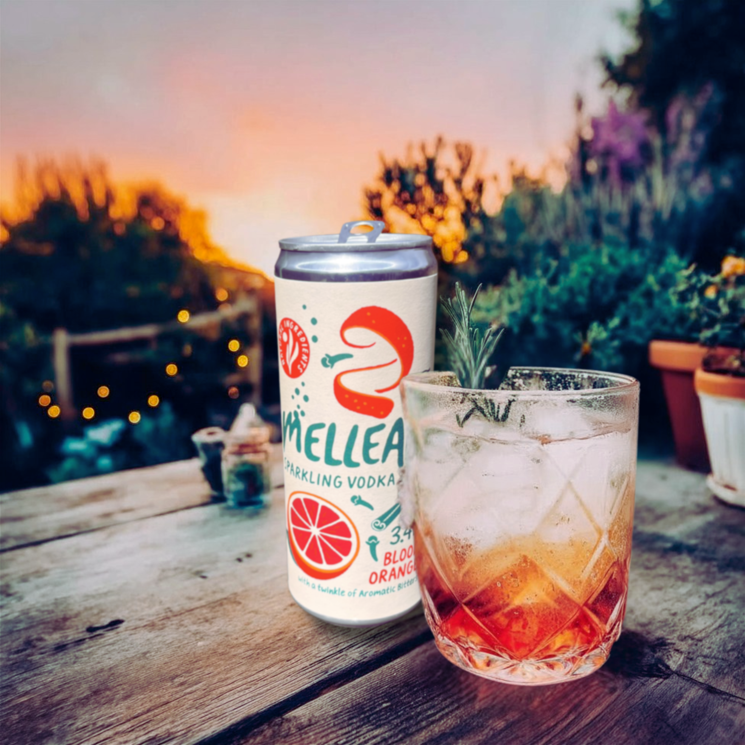 A can of Mellea Sparkling Vodka with blood orange flavor and a glass with a drink and ice on a wooden table outdoors during sunset.