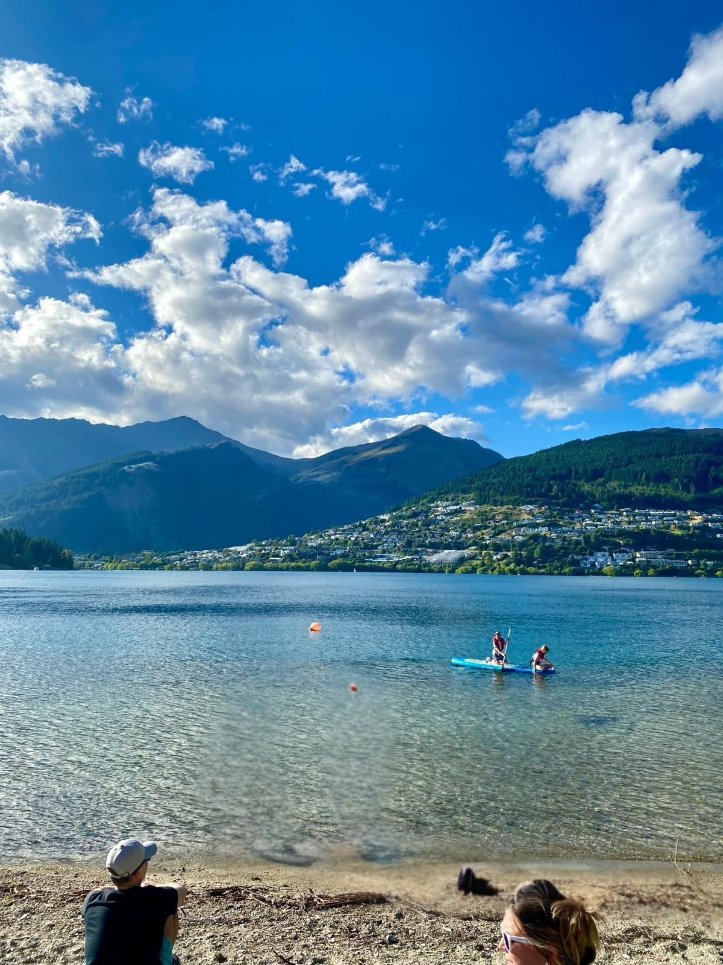 A lakeside scene with a mountain range in the background, two people standing on paddleboards in the water, and two people sitting on the sandy shore in the foreground, one wearing sunglasses and a hat.