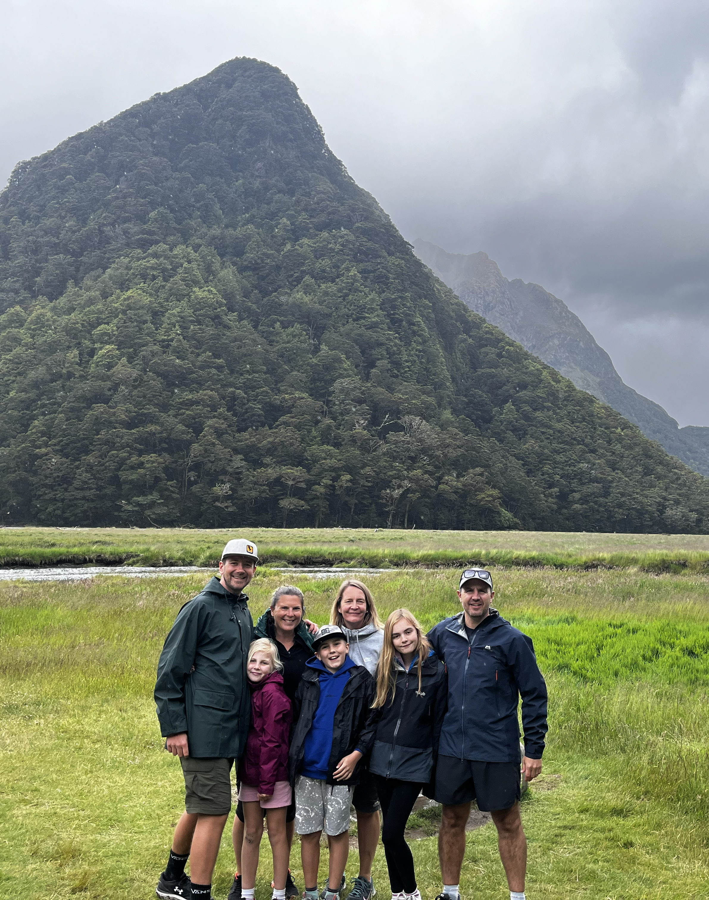 Family of seven posing in front of a large mountain with a grassy field and a narrow river or stream in the background.