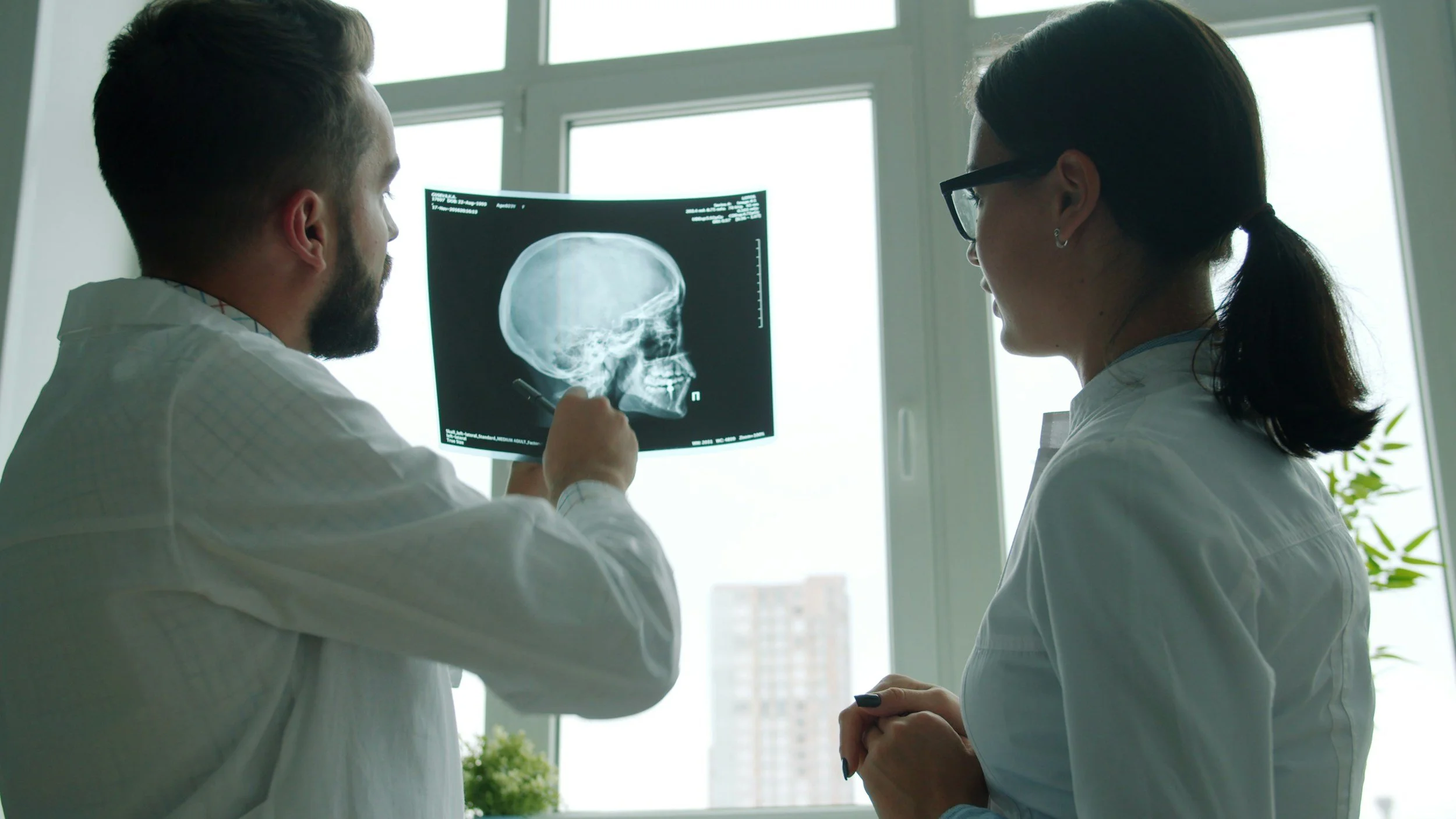 Two medical professionals, a man and a woman, reviewing a brain scan X-ray in a bright room with large windows.