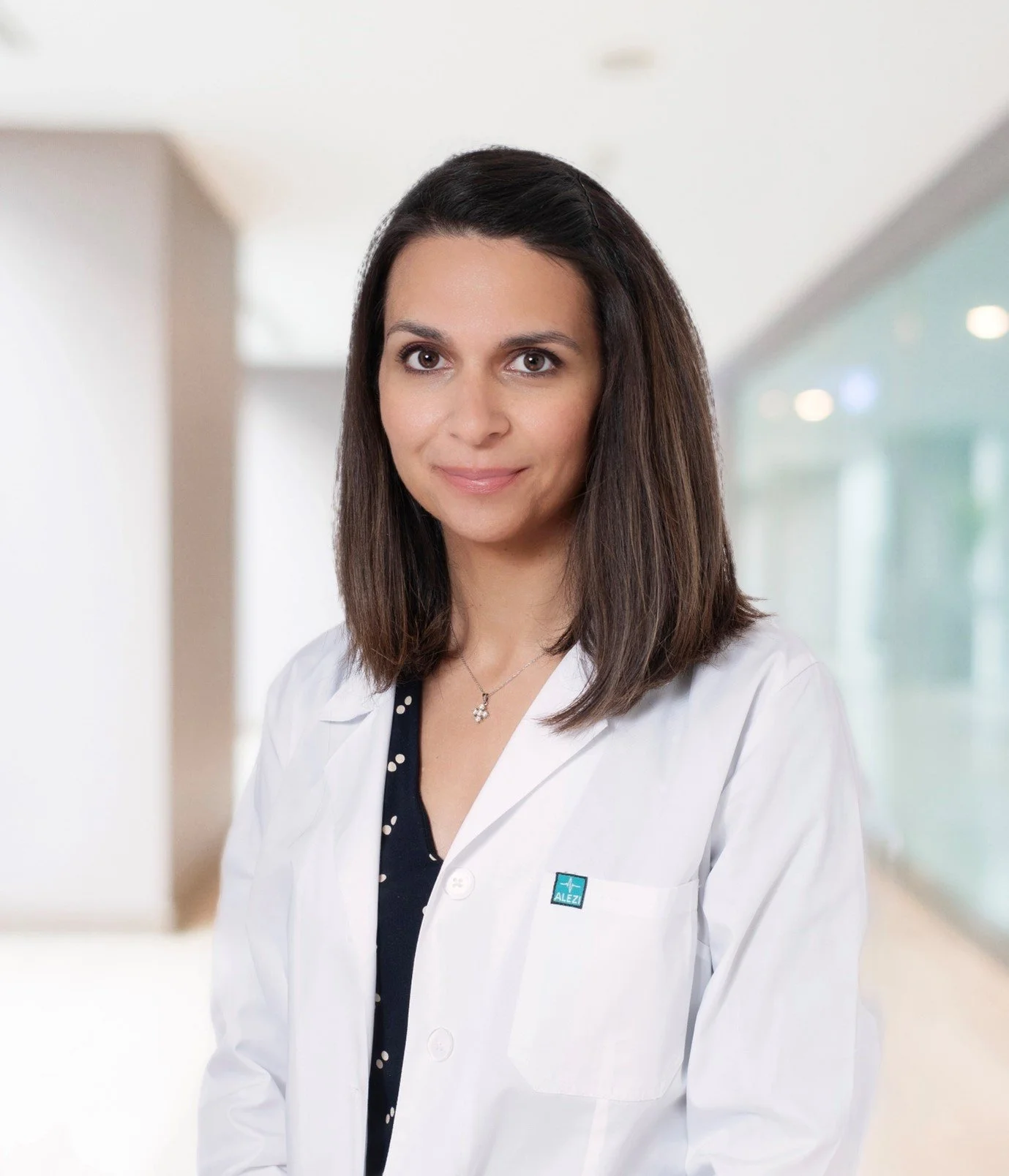 A woman with shoulder-length brown hair wearing a white lab coat and a black blouse, standing in a bright, modern indoor setting.