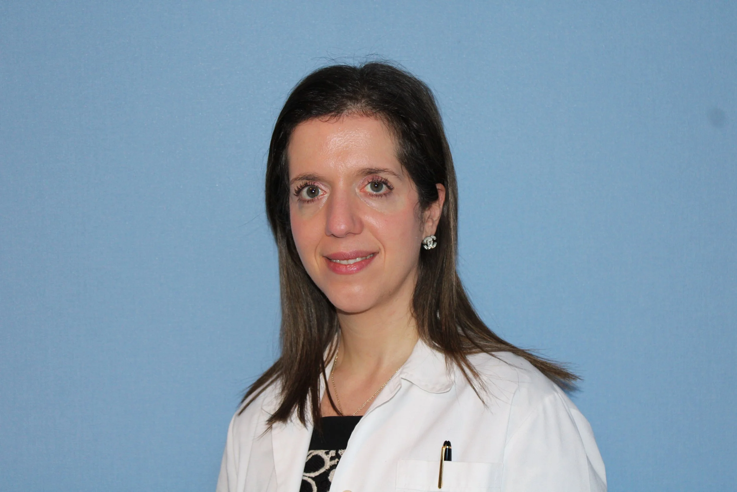 A woman with shoulder-length brown hair, wearing a white coat and earring, stands against a blue background.