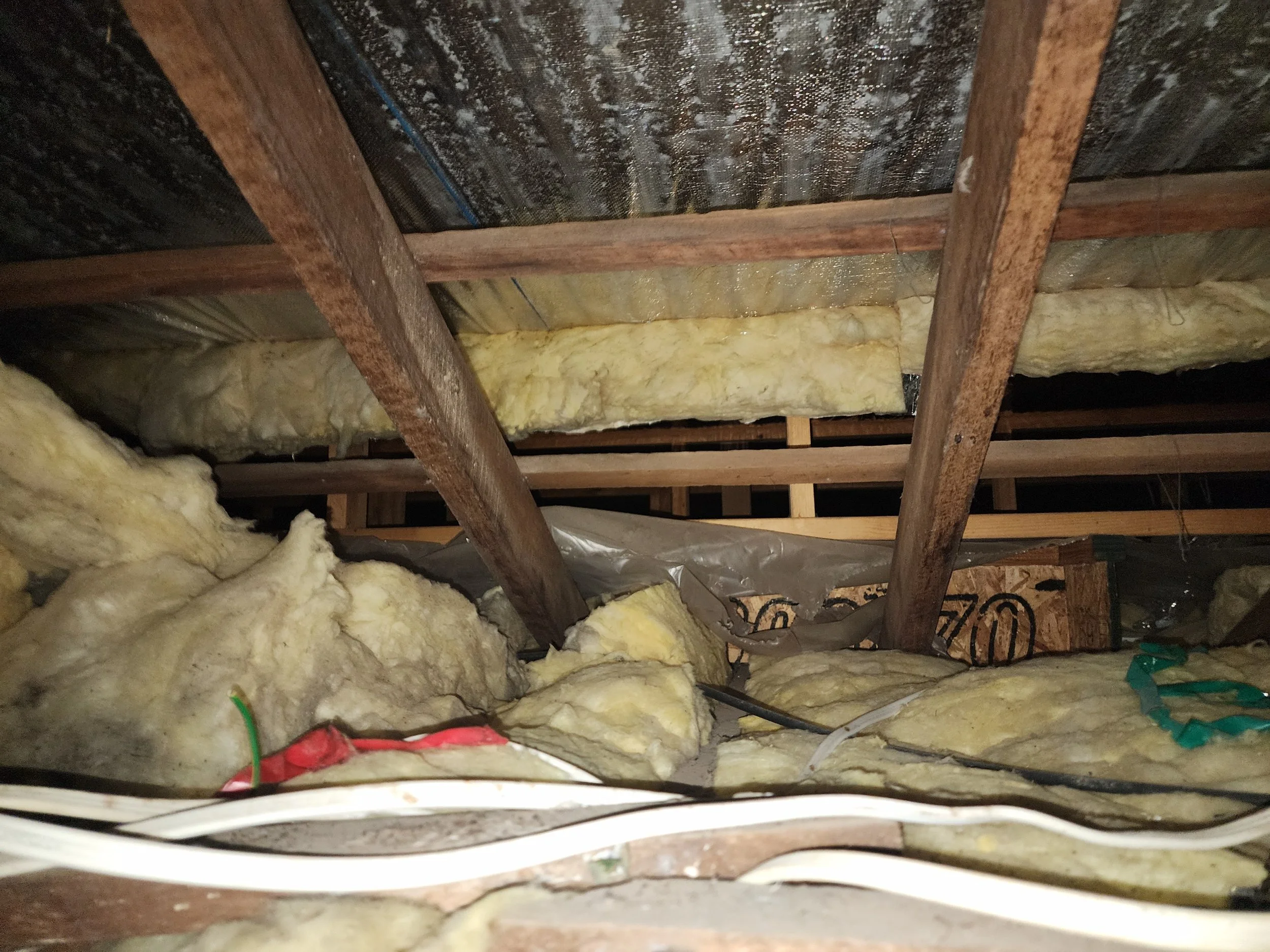Looking up into the attic space showing insulation and wooden framing with some electrical wires.