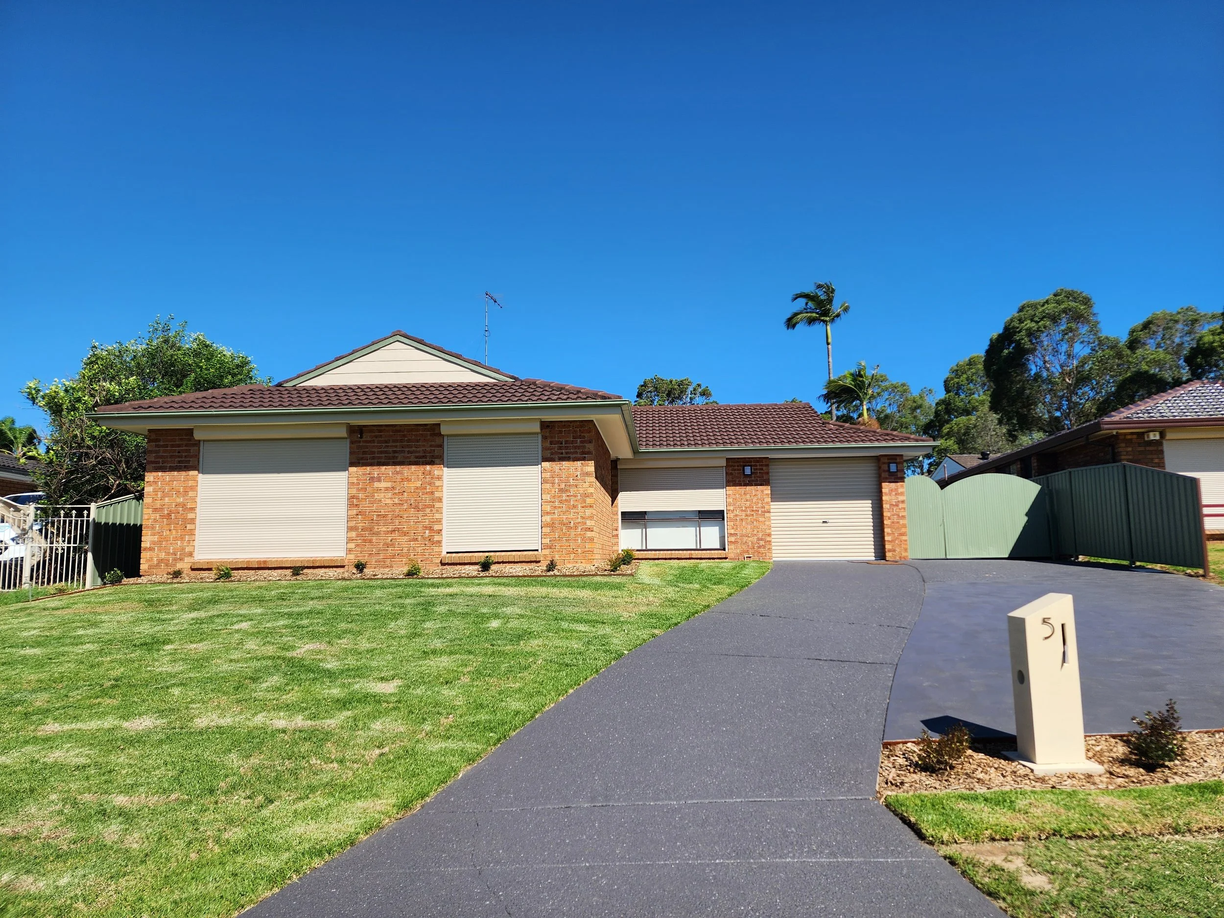 Residential house with a brown tiled roof, brick walls, and a driveway. Front yard has green grass, small bushes, and a sign with the number 51. Sky is clear and blue with a few trees including palm trees.