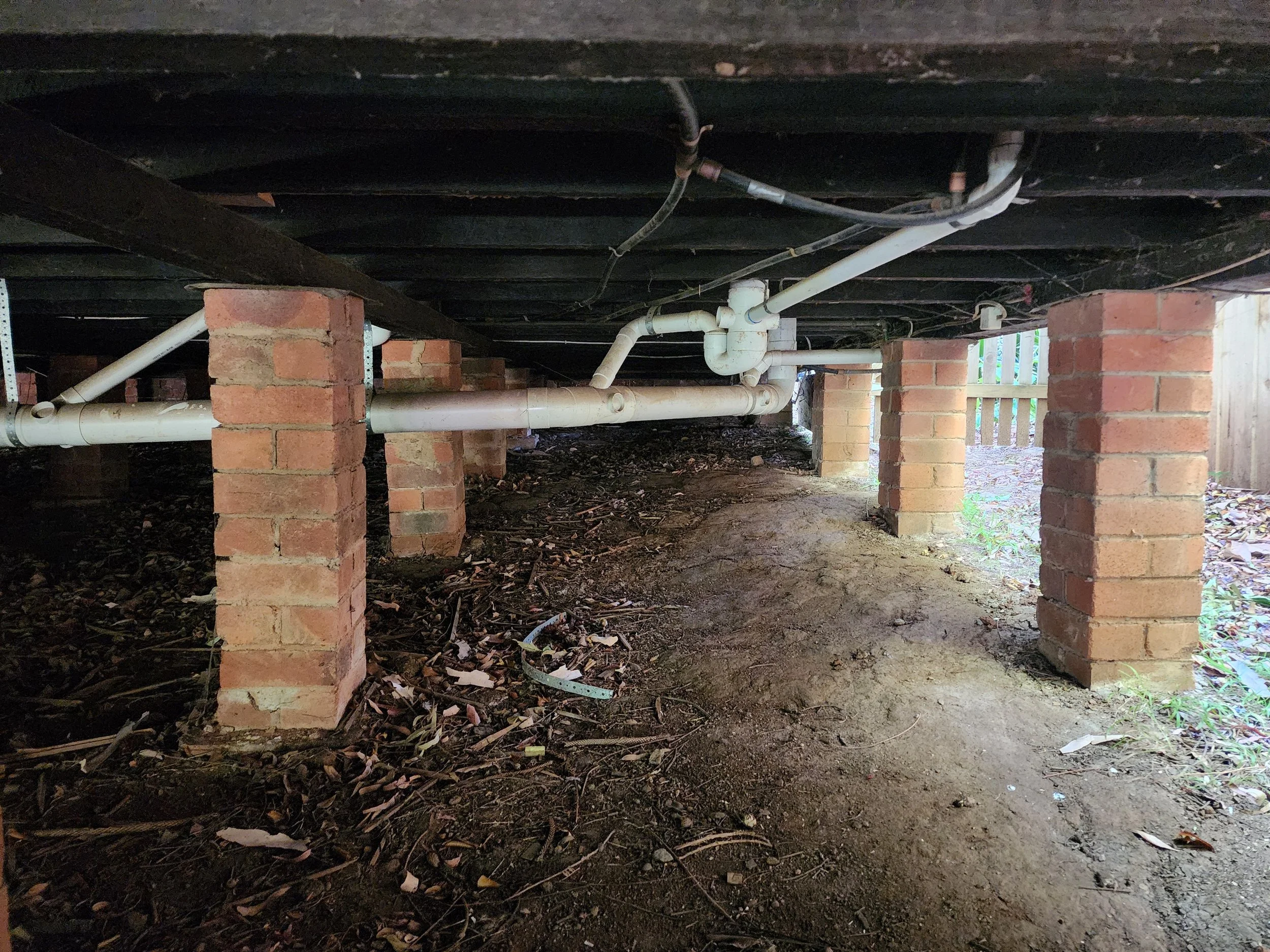Underneath a wooden deck supported by brick pillars, with exposed pipes running through the space.