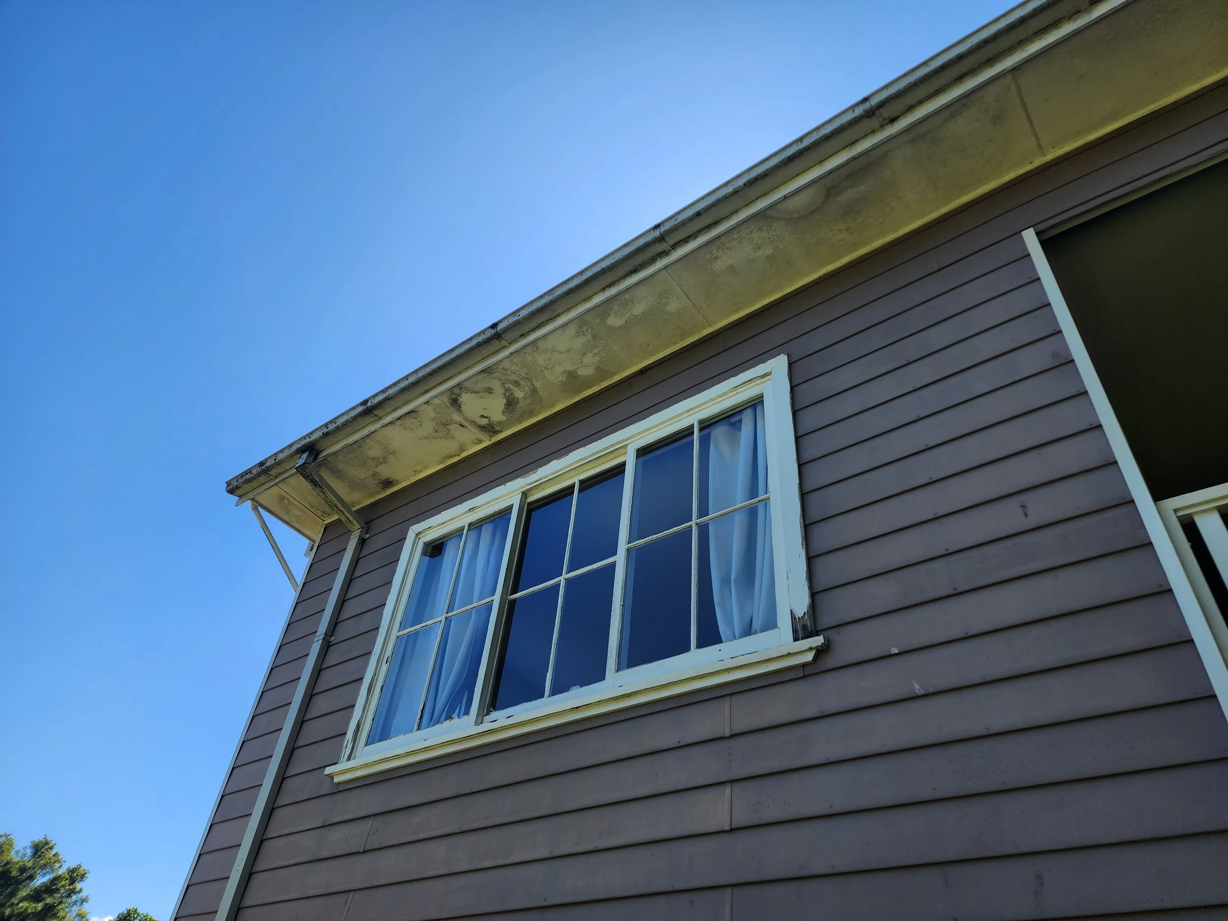 Close-up of the corner of a house with brown siding, white-framed windows, and a yellowish roof edge, against a clear blue sky.