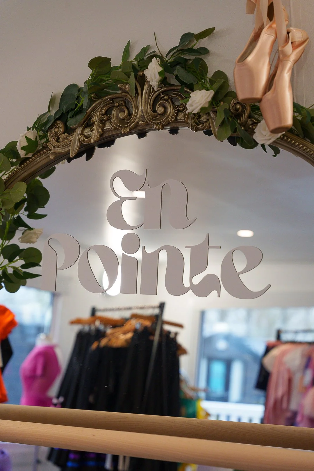 Close-up of a mirror with decorative gold frame and green foliage, with a sign reading 'en pointe' hanging in front. Inside a clothing store with racks of clothes in the background.