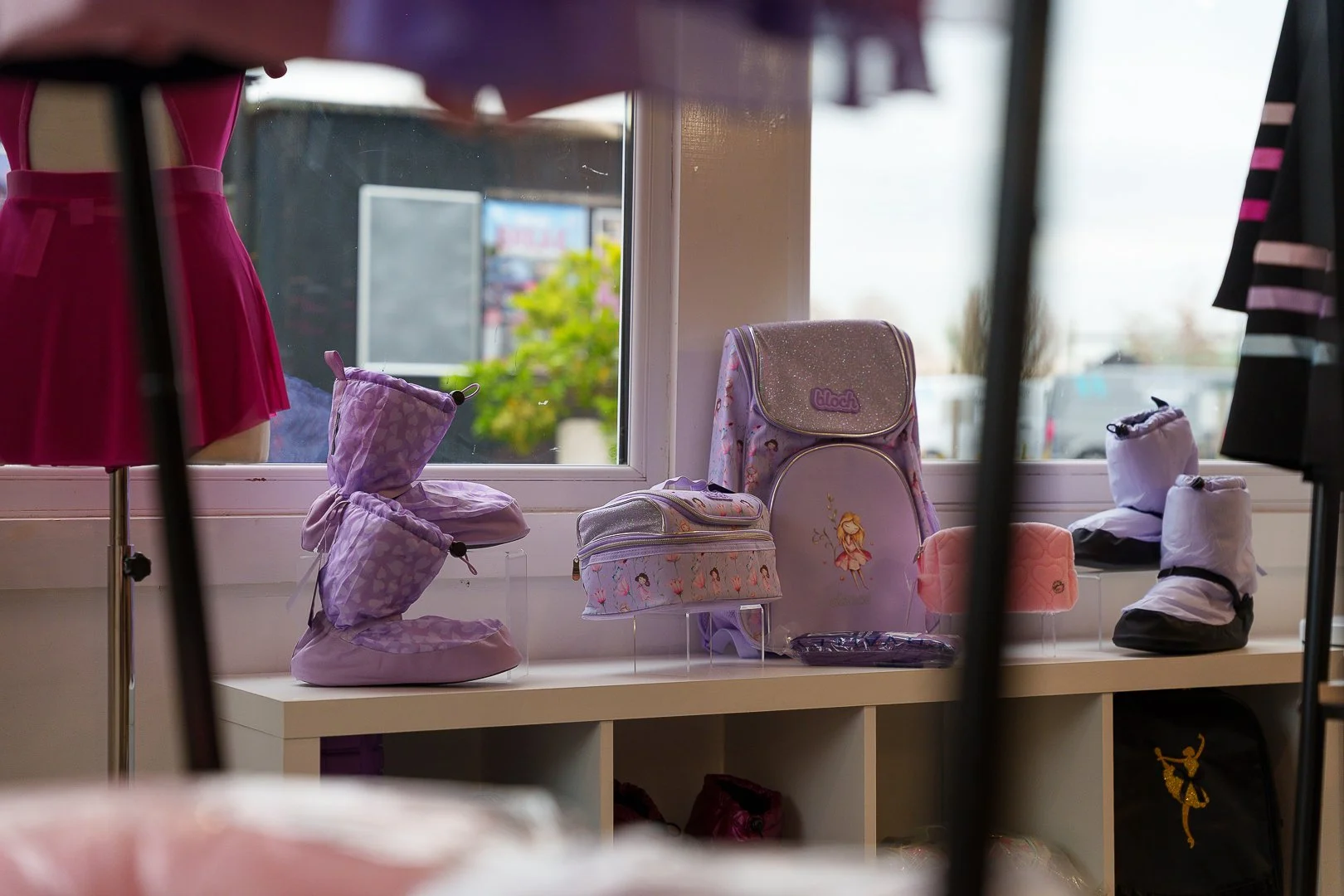Display of purple and pink children's clothing and accessories on a window ledge, including boots, backpacks, and small bags, with a window showing outdoor scenery.