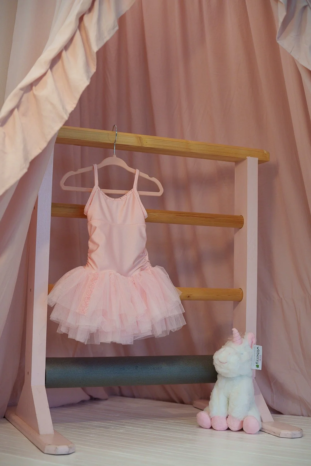 A pink tutu dress hanging on a pink hanger inside a wooden frame, with a pink curtain in the background and a white plush unicorn toy with pink hooves and a pink horn at the base.