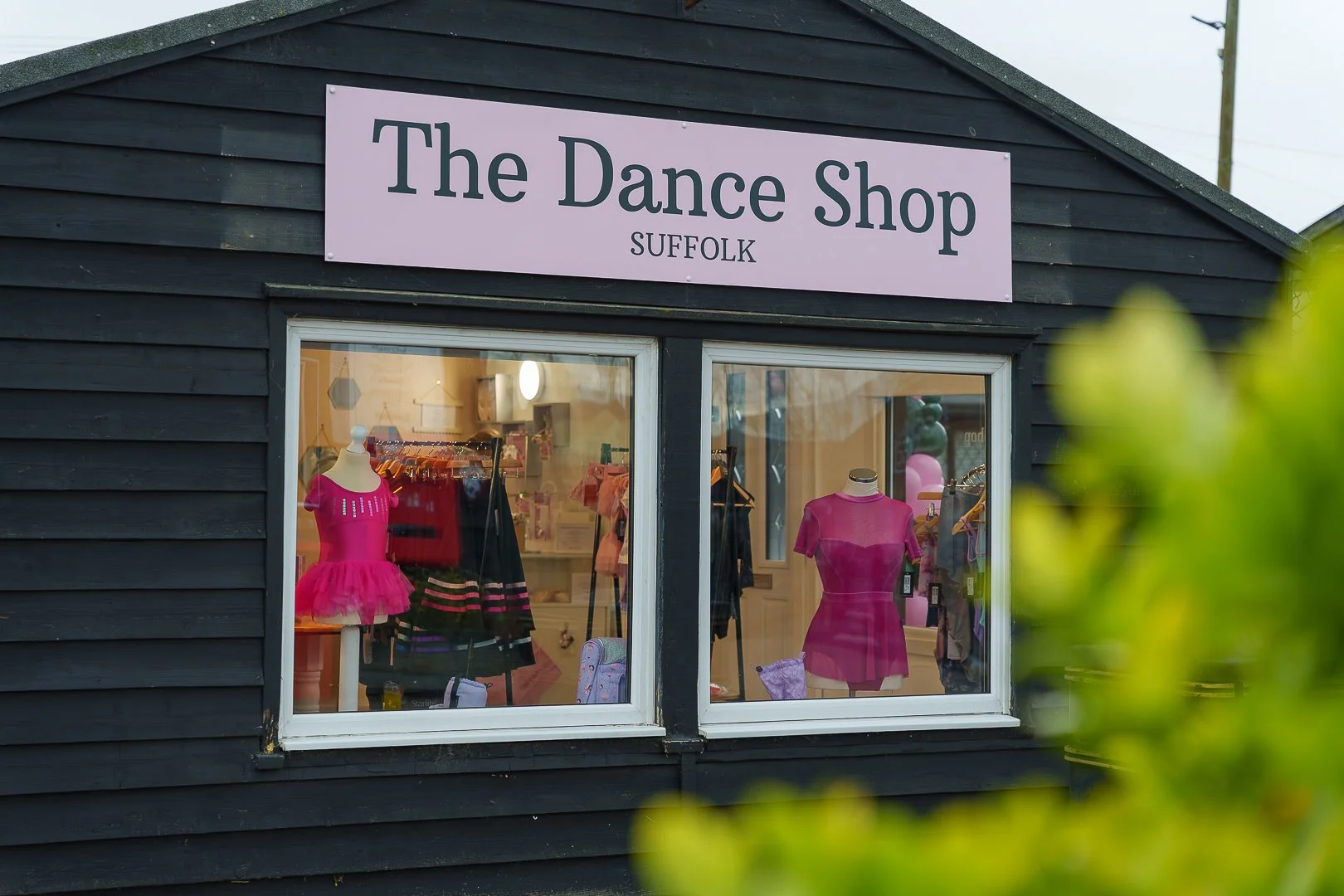 Shopfront of The Dance Shop in Suffolk with pink signage, displaying dance costumes and mannequins dressed in pink and purple dance attire inside.