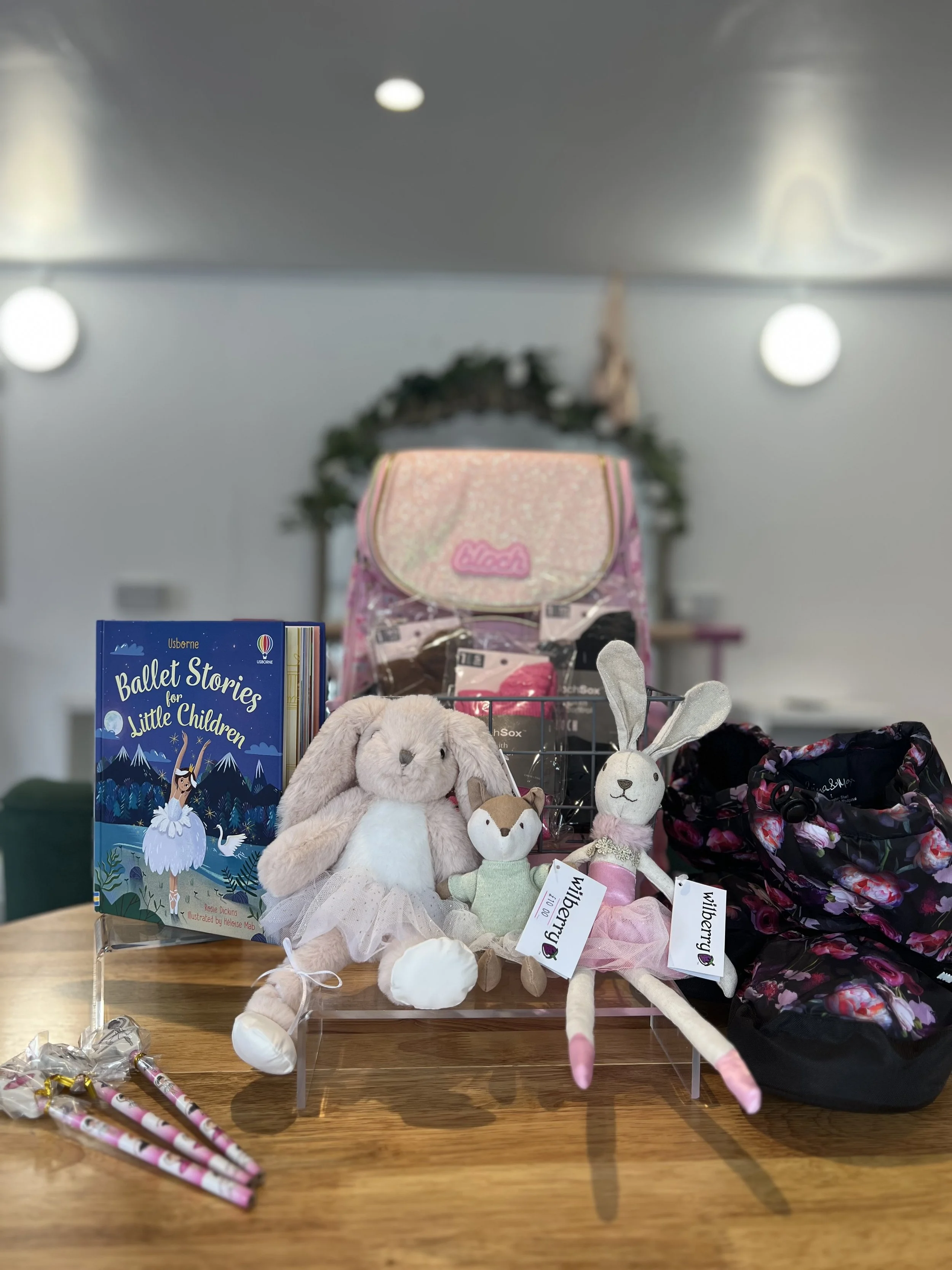 Collection of children's toys and books on a wooden table, including plush rabbits and foxes, a book titled 'Ballet Stories for Little Children,' a pink backpack, and other small items.