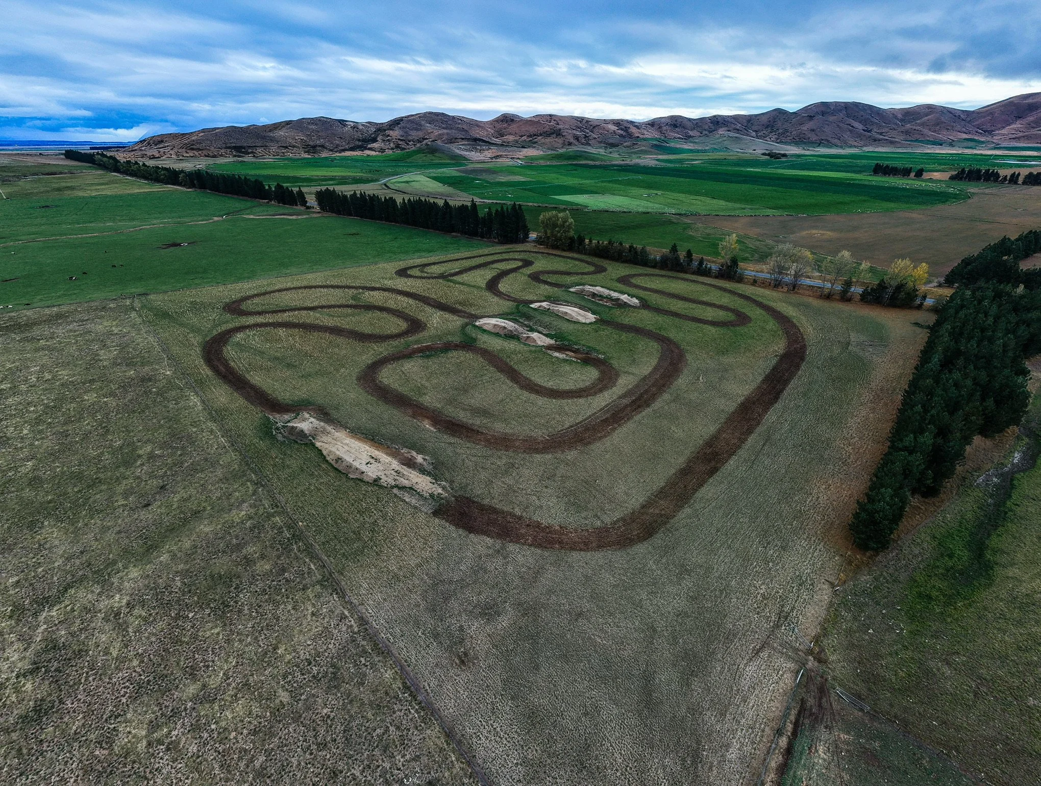 Aerial view of agricultural fields with winding dirt roads, a line of tall dark trees, small patches of tents or structures, and a backdrop of rolling hills under a cloudy sky.